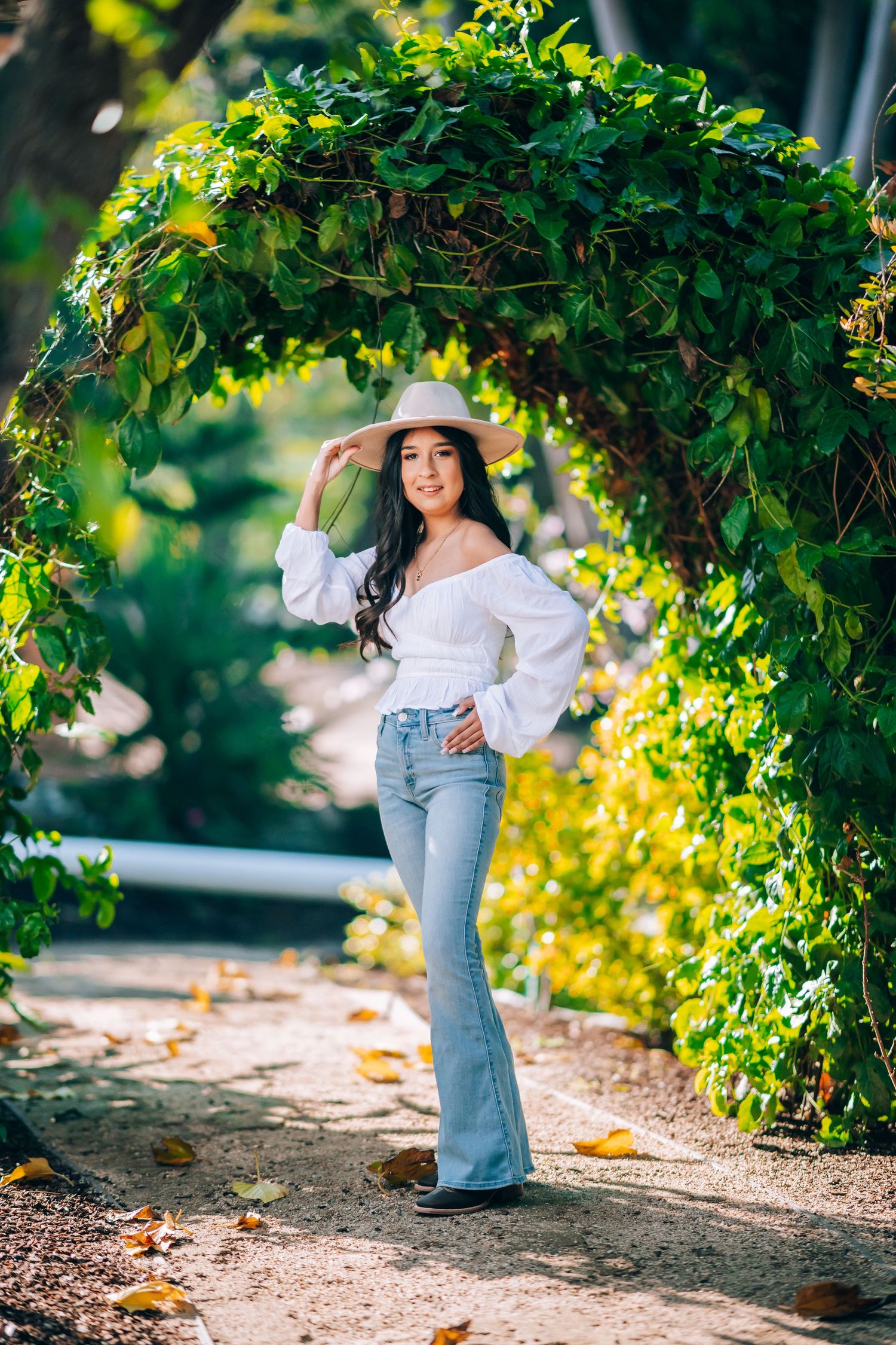 Young woman in white off-shoulder top, flare jeans, and hat poses under a lush green garden archway.