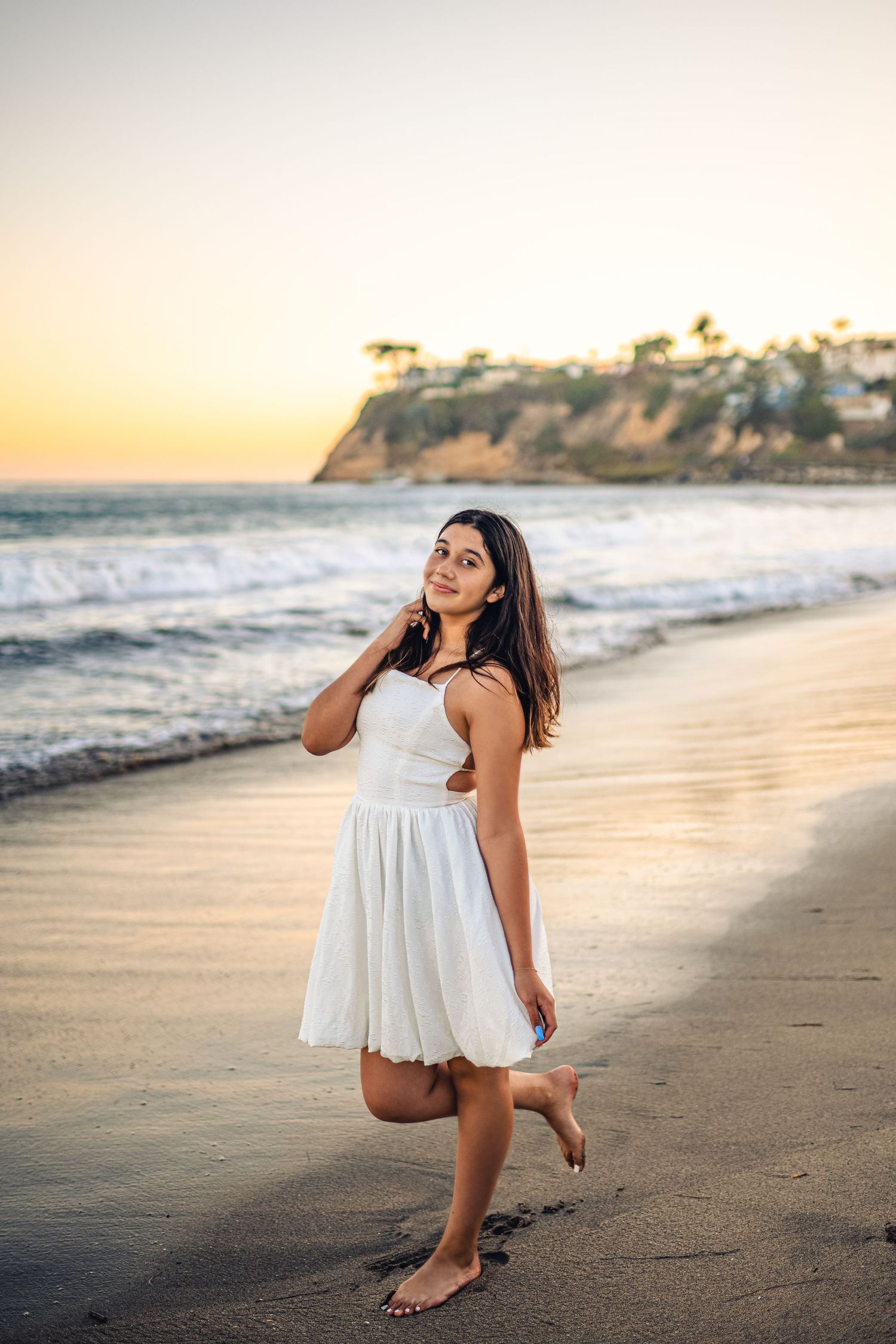 Young woman in white dress poses barefoot on sandy beach at golden sunset with ocean waves and coastal cliffs behind her.