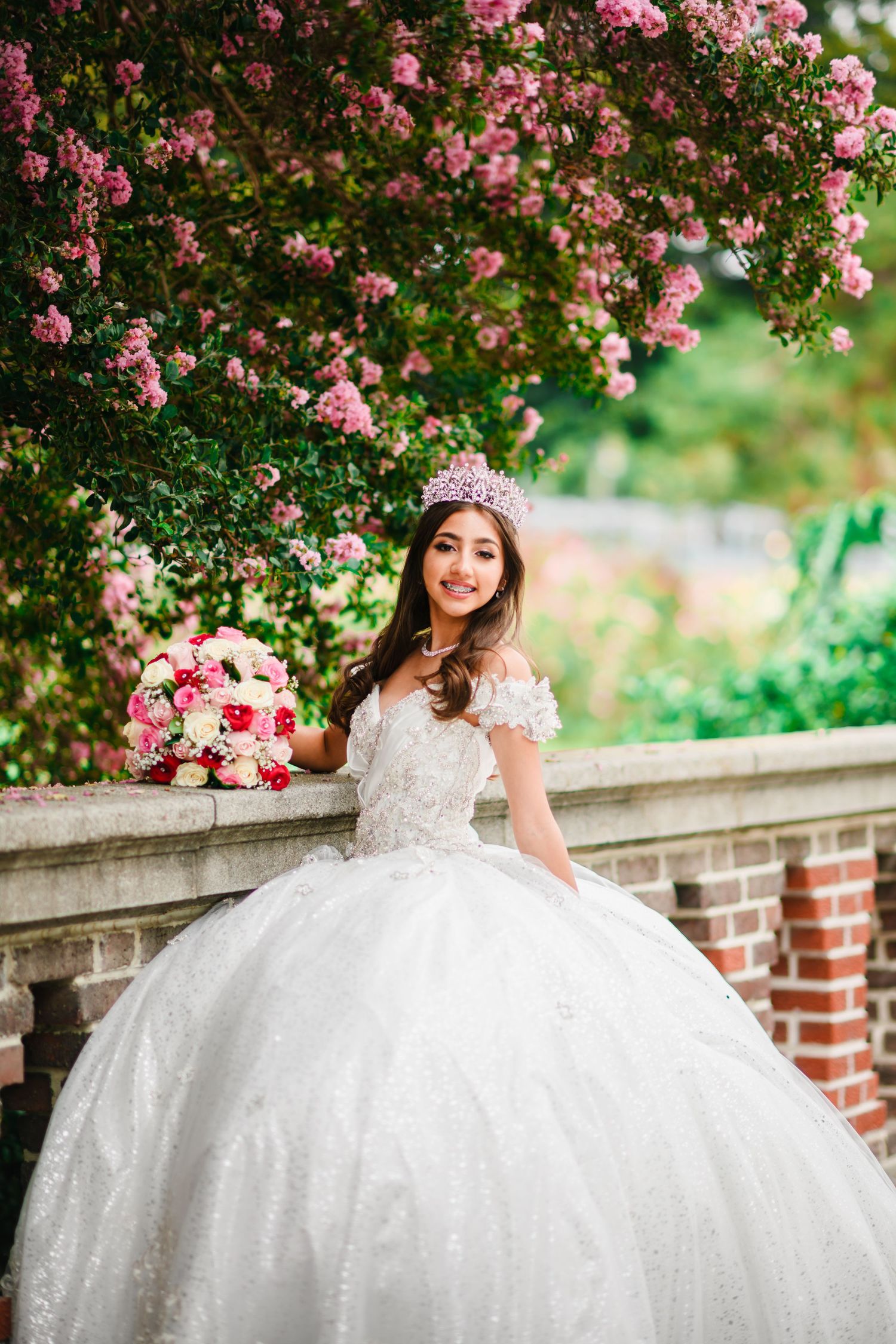 Young bride in white ball gown with tiara holds pink bouquet near blooming rose garden balustrade.
