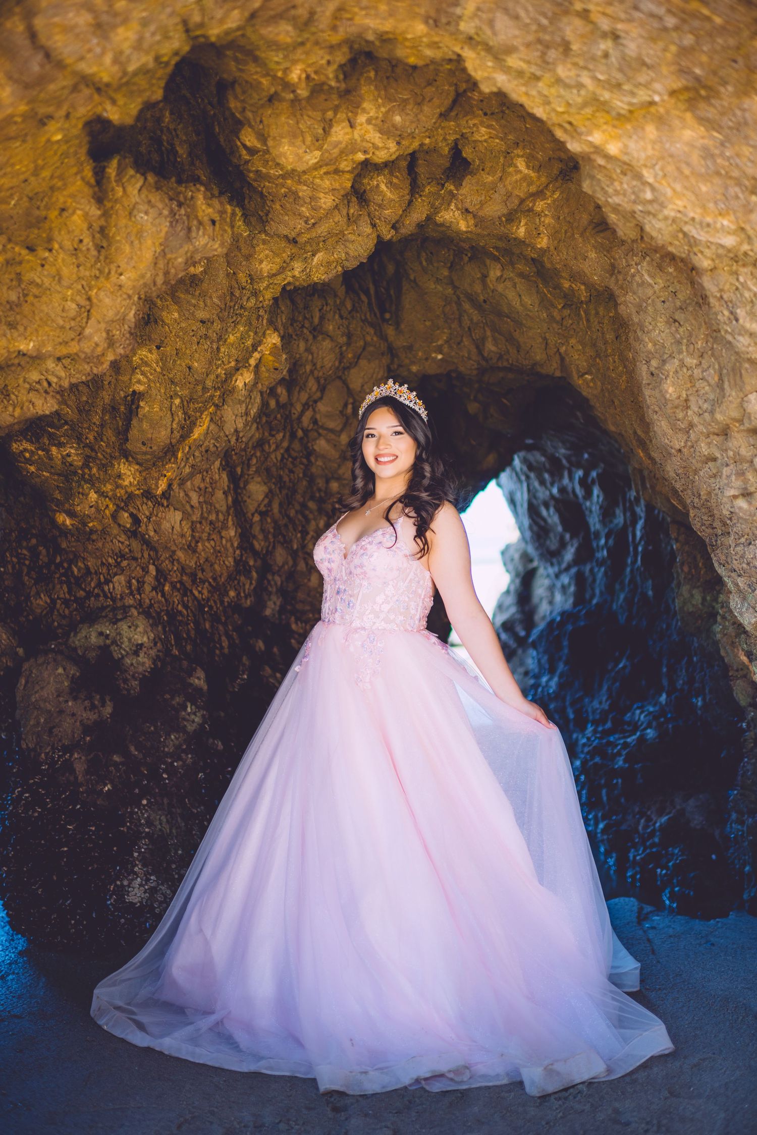 A pink ball gown with glittering details photographed inside a dramatic cave setting with golden rock walls.