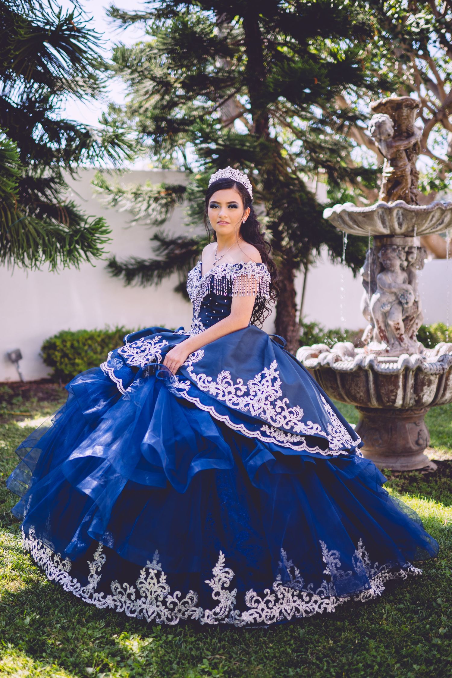 Dramatic navy blue quinceañera ball gown with white lace appliques photographed in a garden setting.
