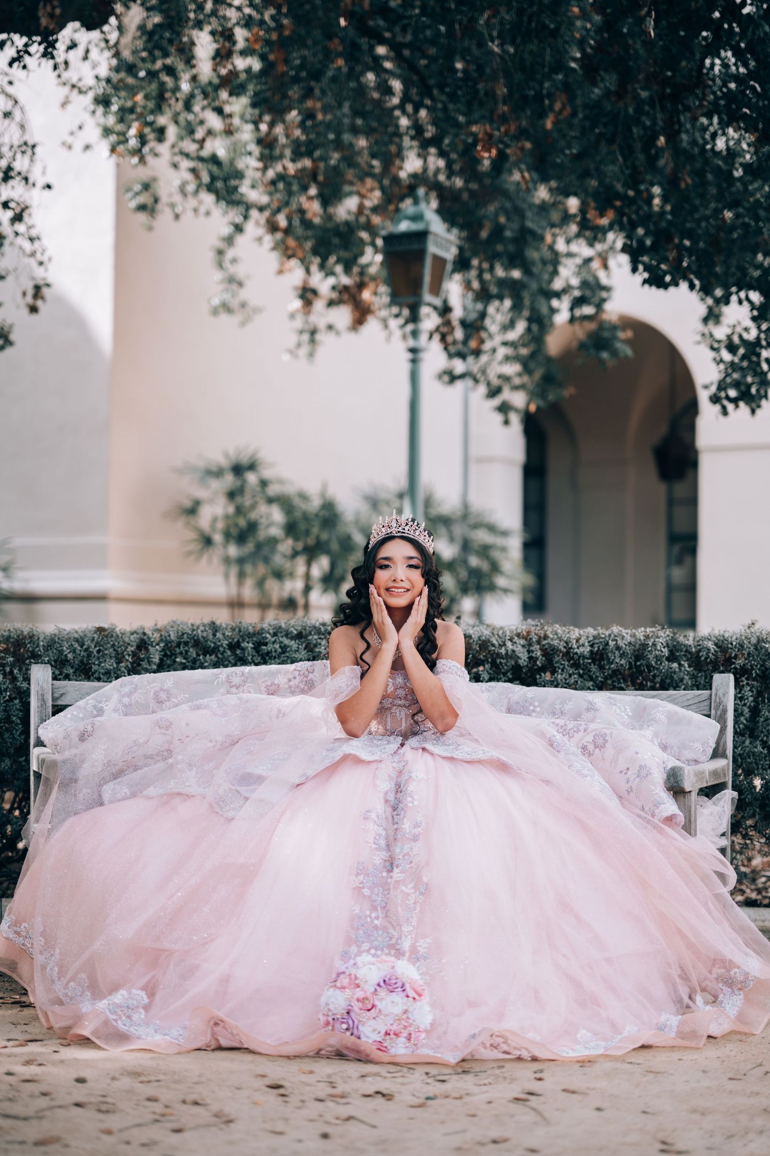 Young woman in pink ball gown and tiara sits on bench, smiling with hands on cheeks, quinceañera celebration.