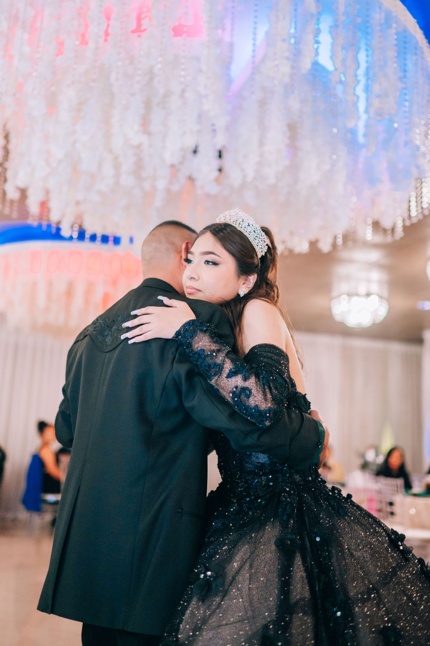 A couple embraces on the dance floor beneath sparkling decorations at an elegant ballroom celebration.