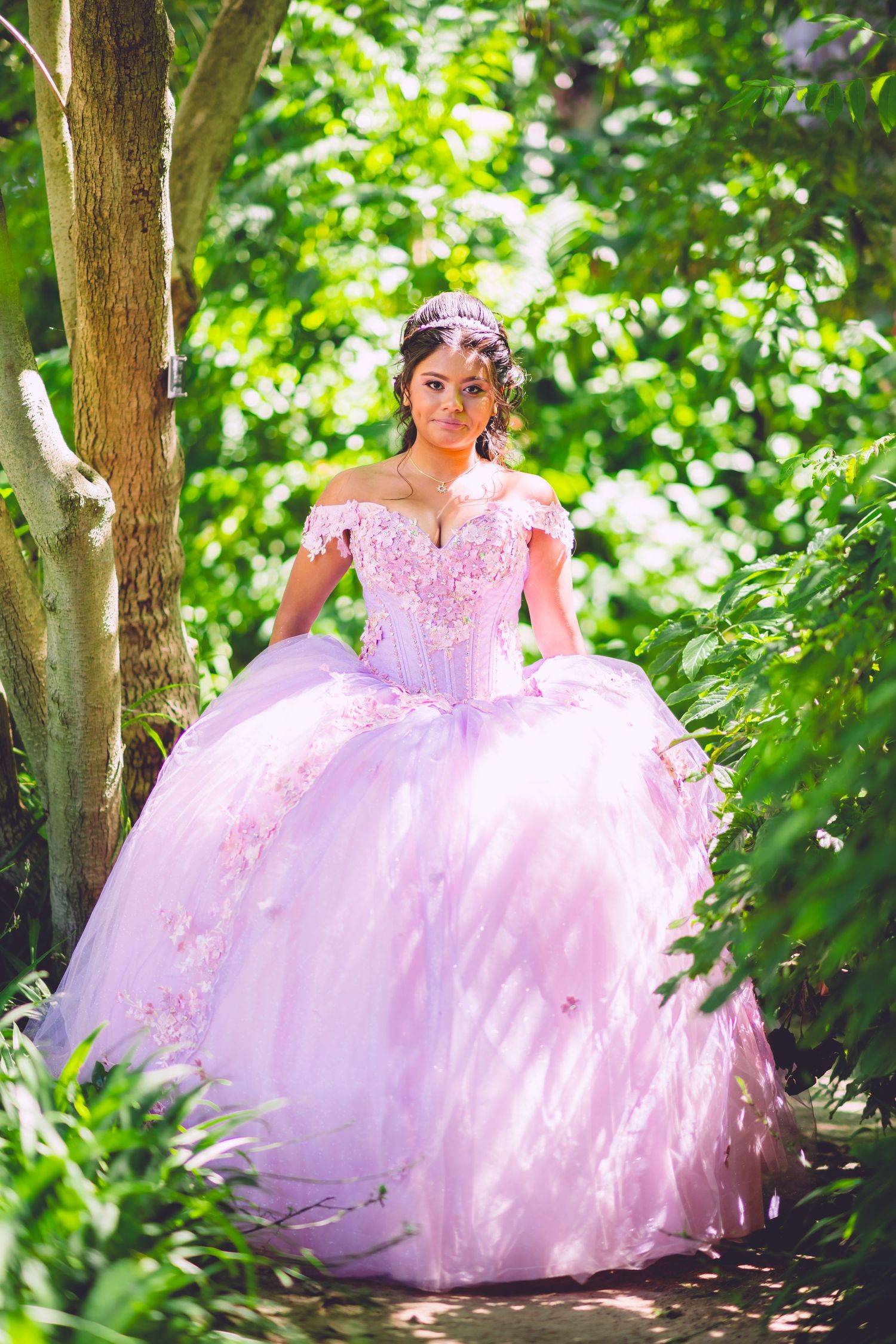 A beautiful pink ball gown with lace details sparkles in dappled sunlight among green garden foliage.
