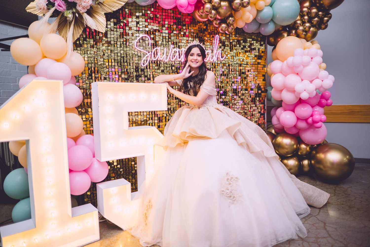 A young woman in a white ballgown poses beside illuminated number 15 at a quinceañera celebration with colorful balloons.