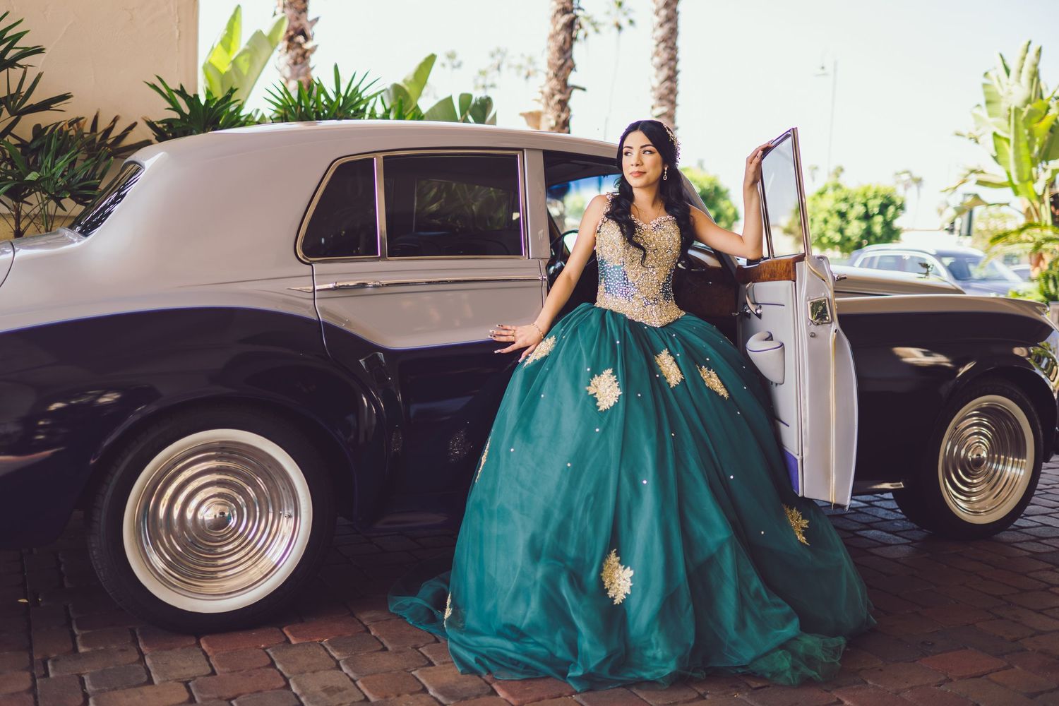 A person in an emerald green ball gown poses elegantly next to vintage cars in a tropical outdoor setting.