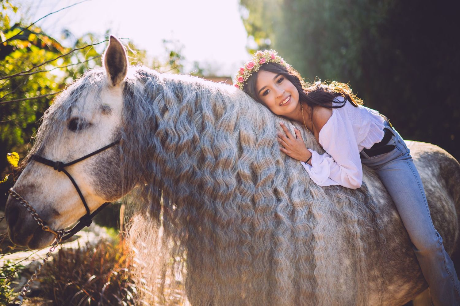 A smiling girl with a flower crown leans affectionately on a white horse with curly mane outdoors.
