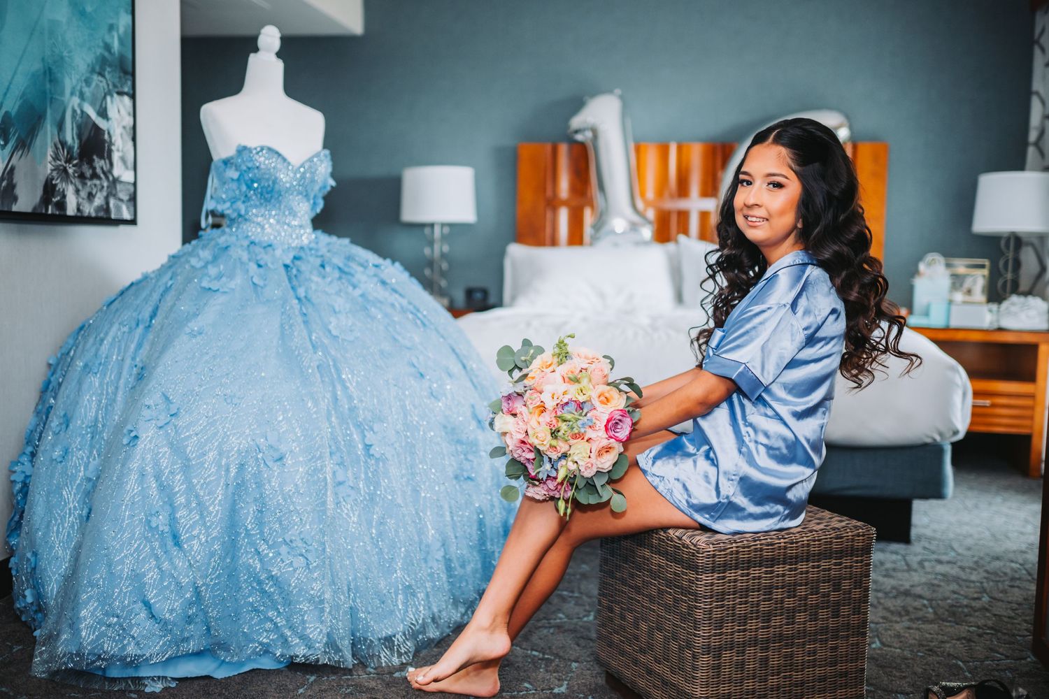 A light blue ballgown with intricate beading is displayed next to someone holding a pink floral bouquet.