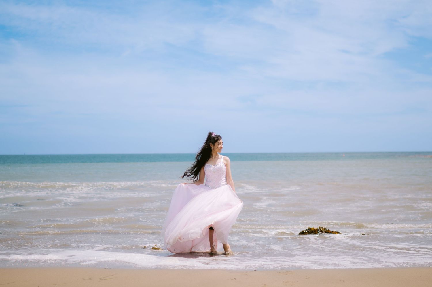 A woman in a flowing pink ball gown stands barefoot at the ocean shoreline on a sandy beach under a blue sky.
