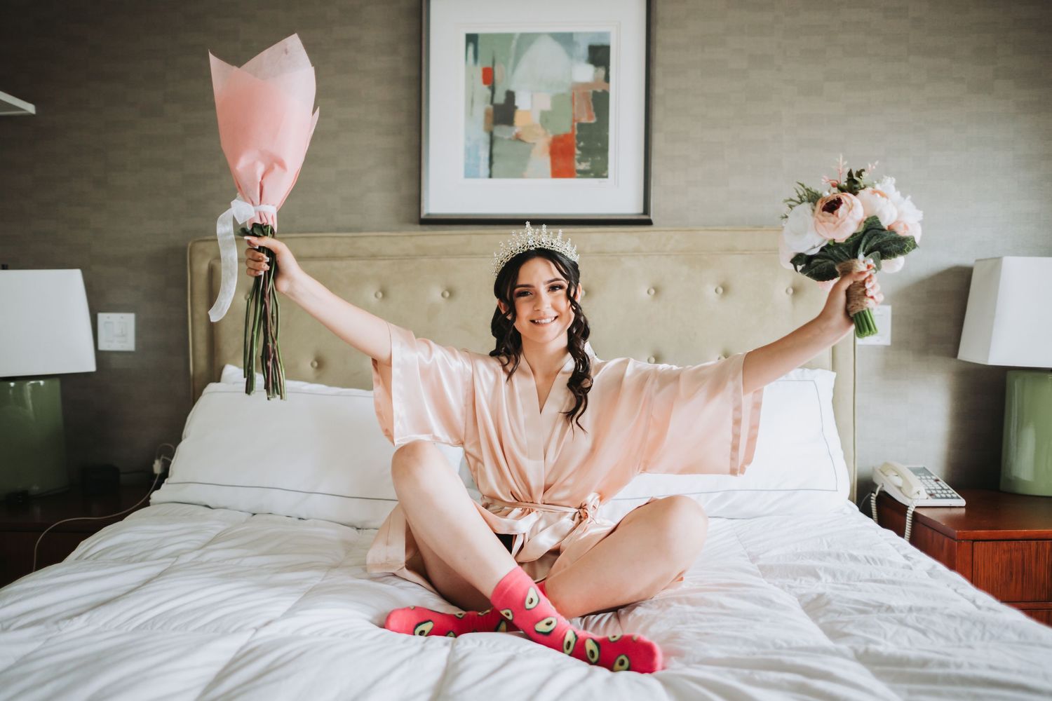 Smiling bride in pink robe sits cross-legged on hotel bed, holding bouquets aloft, wearing crown and red bridal socks.