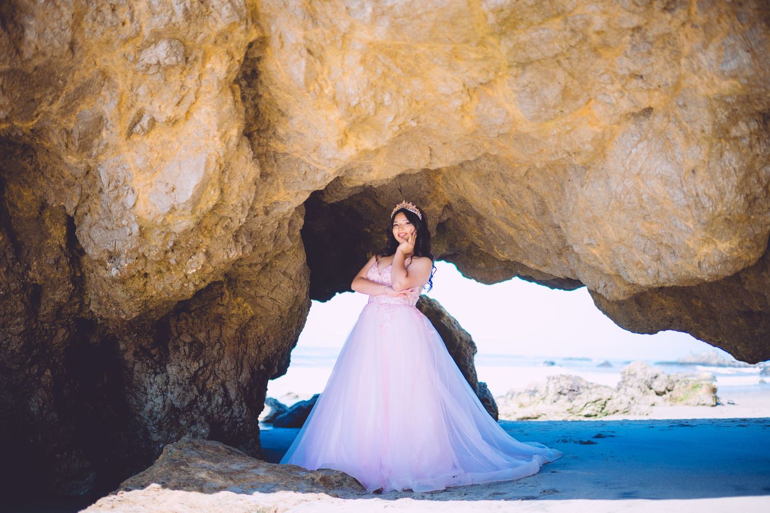 Woman in pink ball gown poses gracefully inside a rocky coastal cave arch with ocean views behind her.