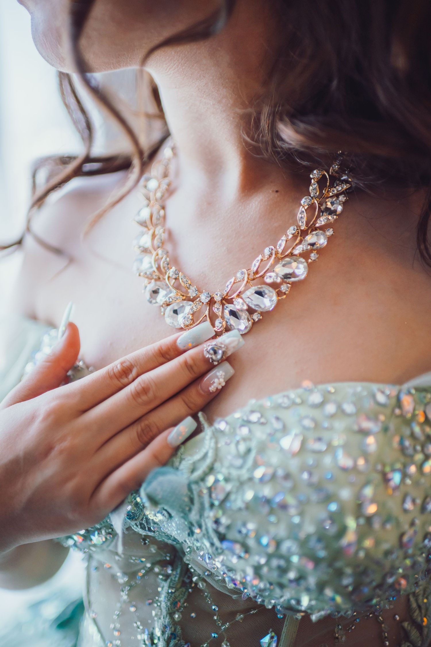 Bride wearing elegant crystal necklace with leaf design, touching jewelry against sparkling mint green beaded gown.