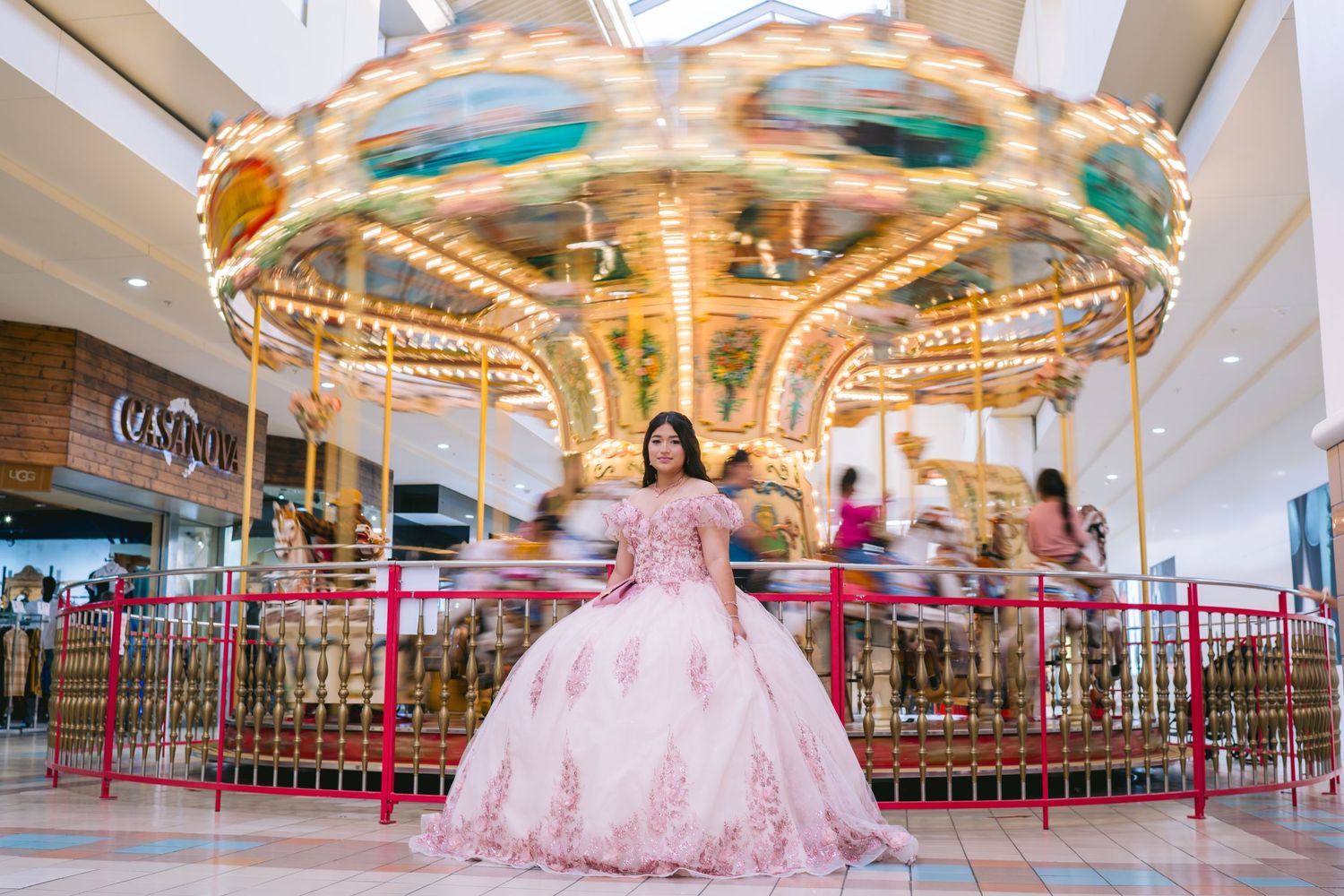 A pink quinceañera dress sparkles in front of an illuminated carousel at a shopping mall.