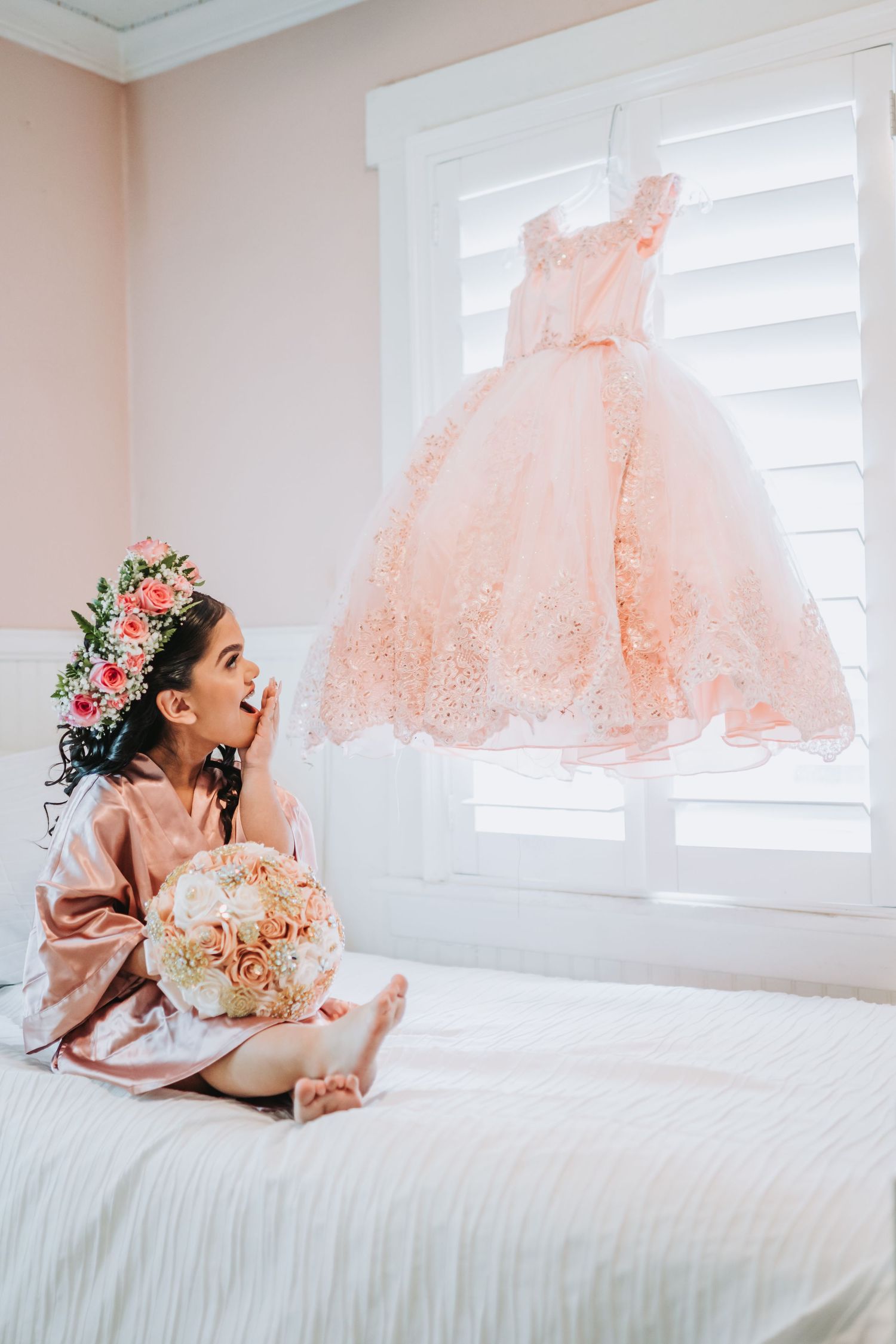 Young figure in pink robe gazes up at a peach tulle dress hanging by window while holding floral bouquet.