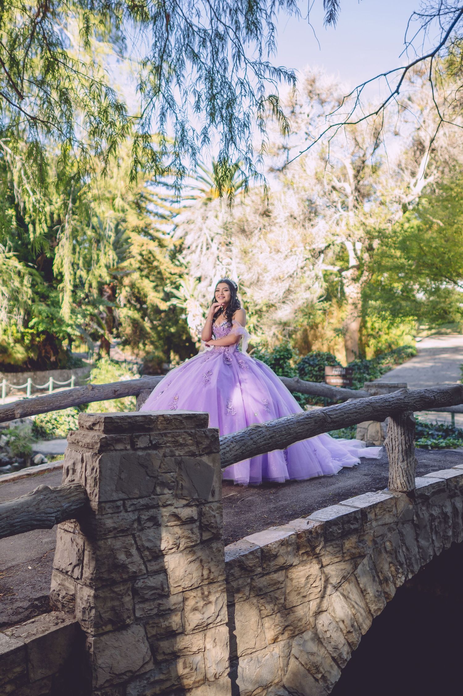 A flowing purple gown creates a dreamy scene against a rustic stone wall backdrop in a sunlit garden setting.