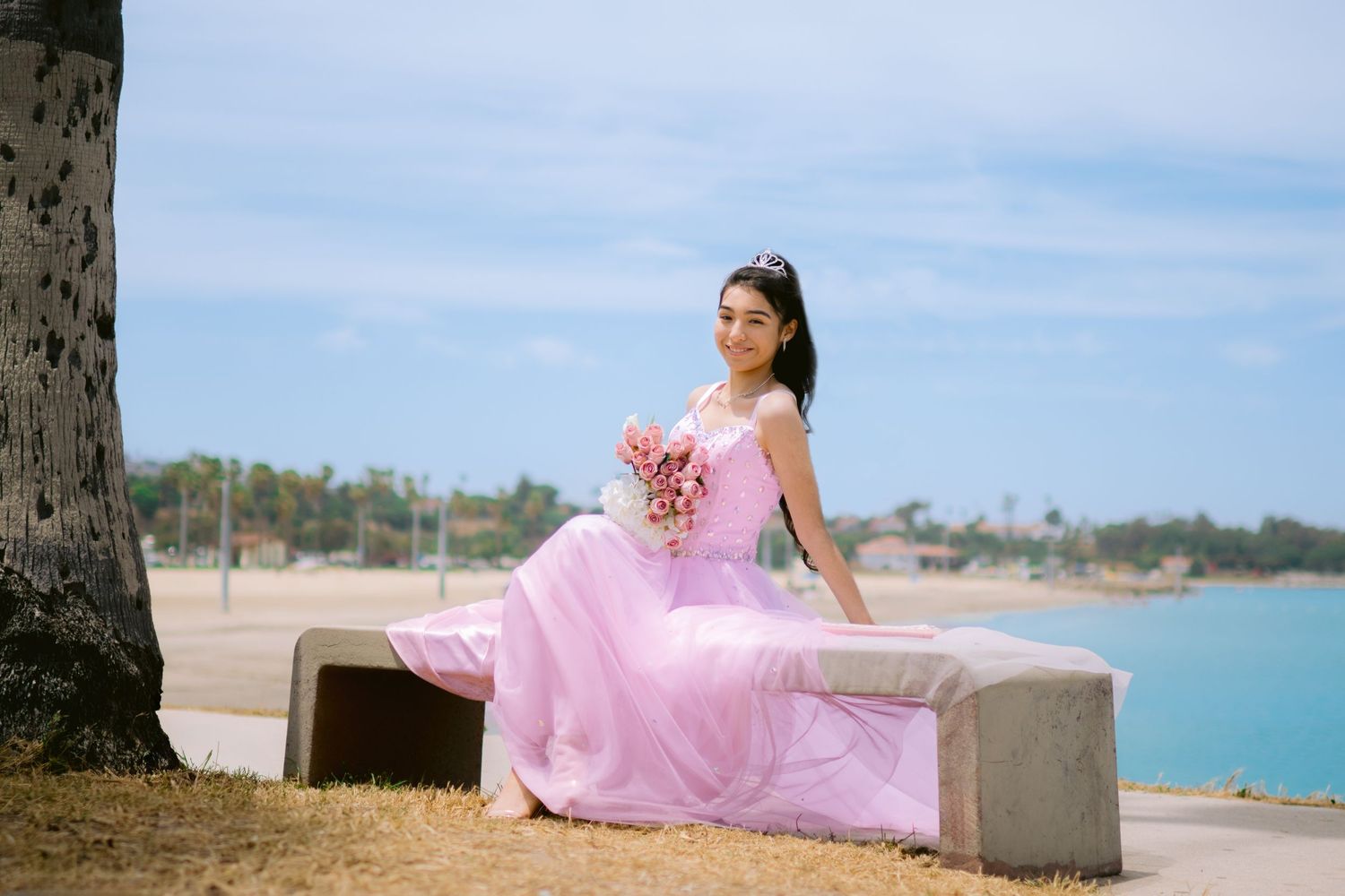 A person in a flowing pink gown poses gracefully on a beach with the ocean and palm trees in the background.