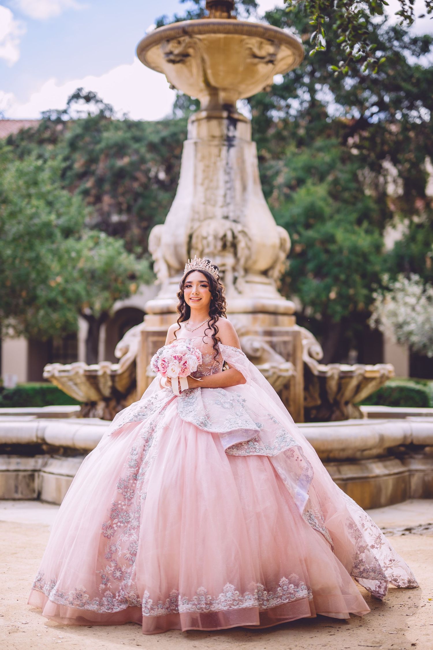 Young woman in pink quinceañera ball gown with floral lace holds bouquet before ornate stone fountain outdoors.