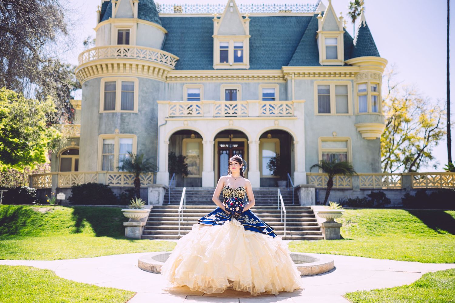 Young woman in a navy and gold ball gown quinceañera dress poses in front of a Victorian mansion.