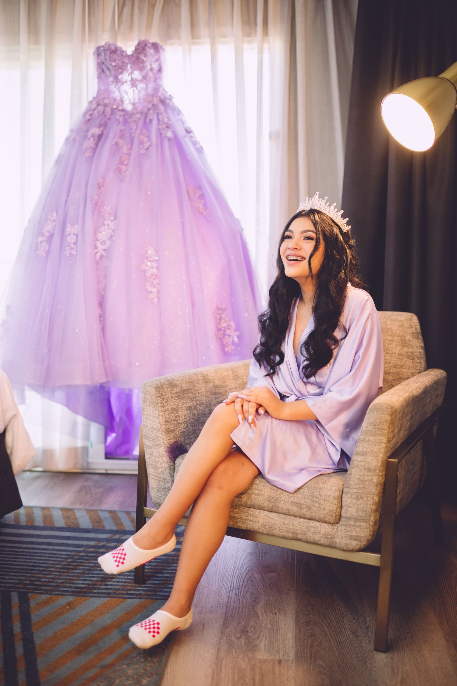 A lavender ball gown hangs beside a plush chair where someone sits in a robe wearing slippers and a tiara.