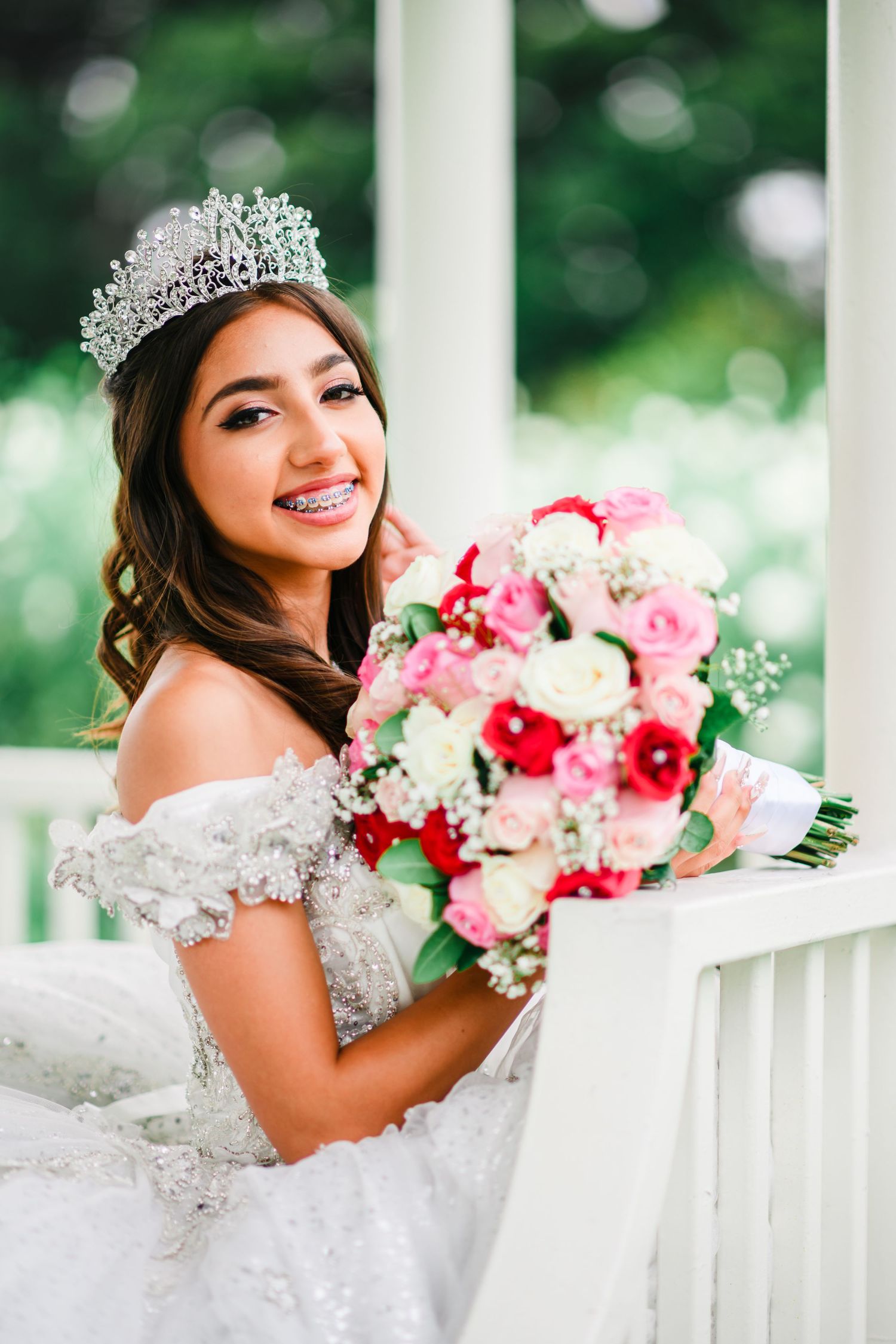 A bride in a white lace dress and crystal tiara holds a pink and red rose bouquet while sitting on a white porch railing.