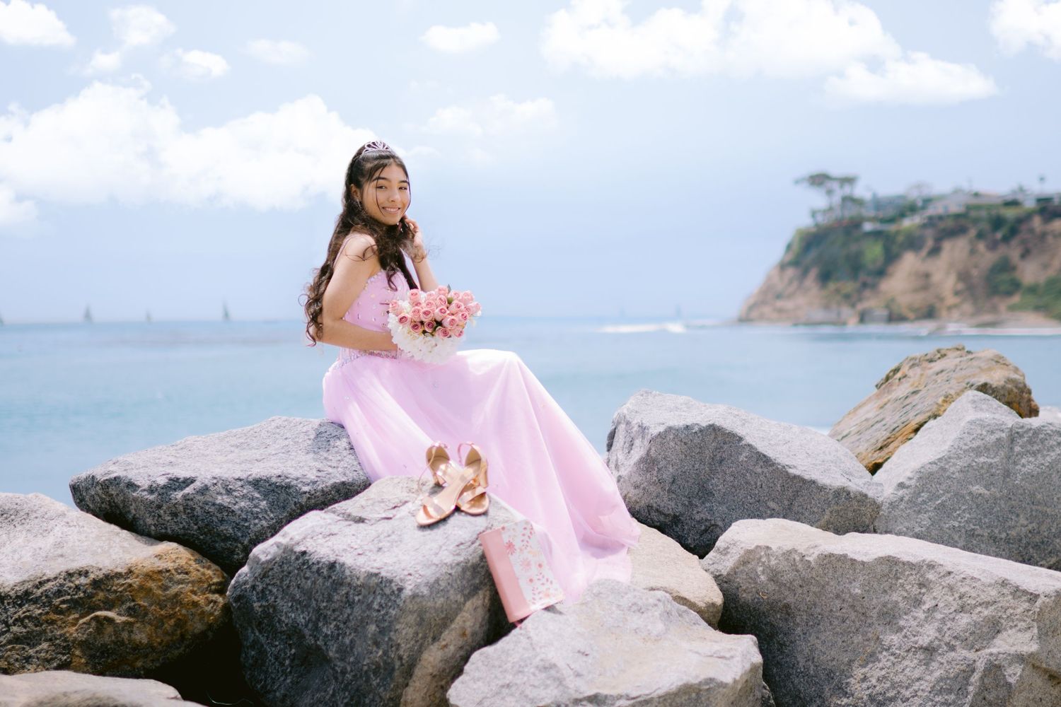 A person in a flowing pink dress poses gracefully on coastal rocks during sunset by the ocean.