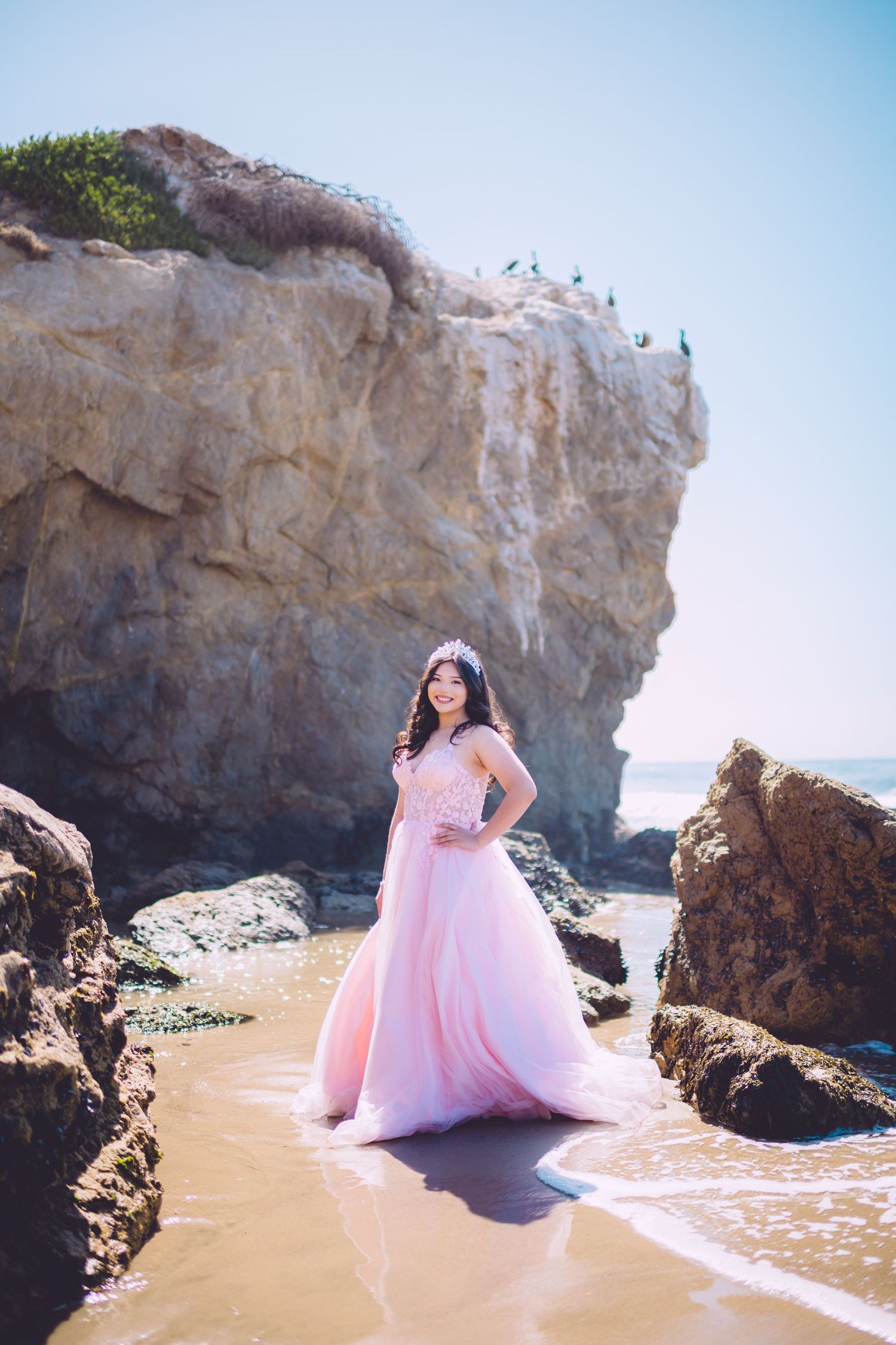 A person in a flowing pink ball gown poses gracefully on a rocky beach with dramatic coastal cliffs in the background.