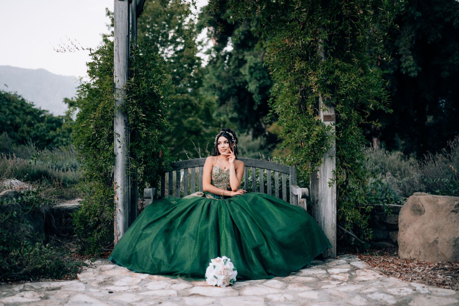 Young woman in elegant emerald green ball gown seated on a bench in a lush garden with a white bouquet nearby.