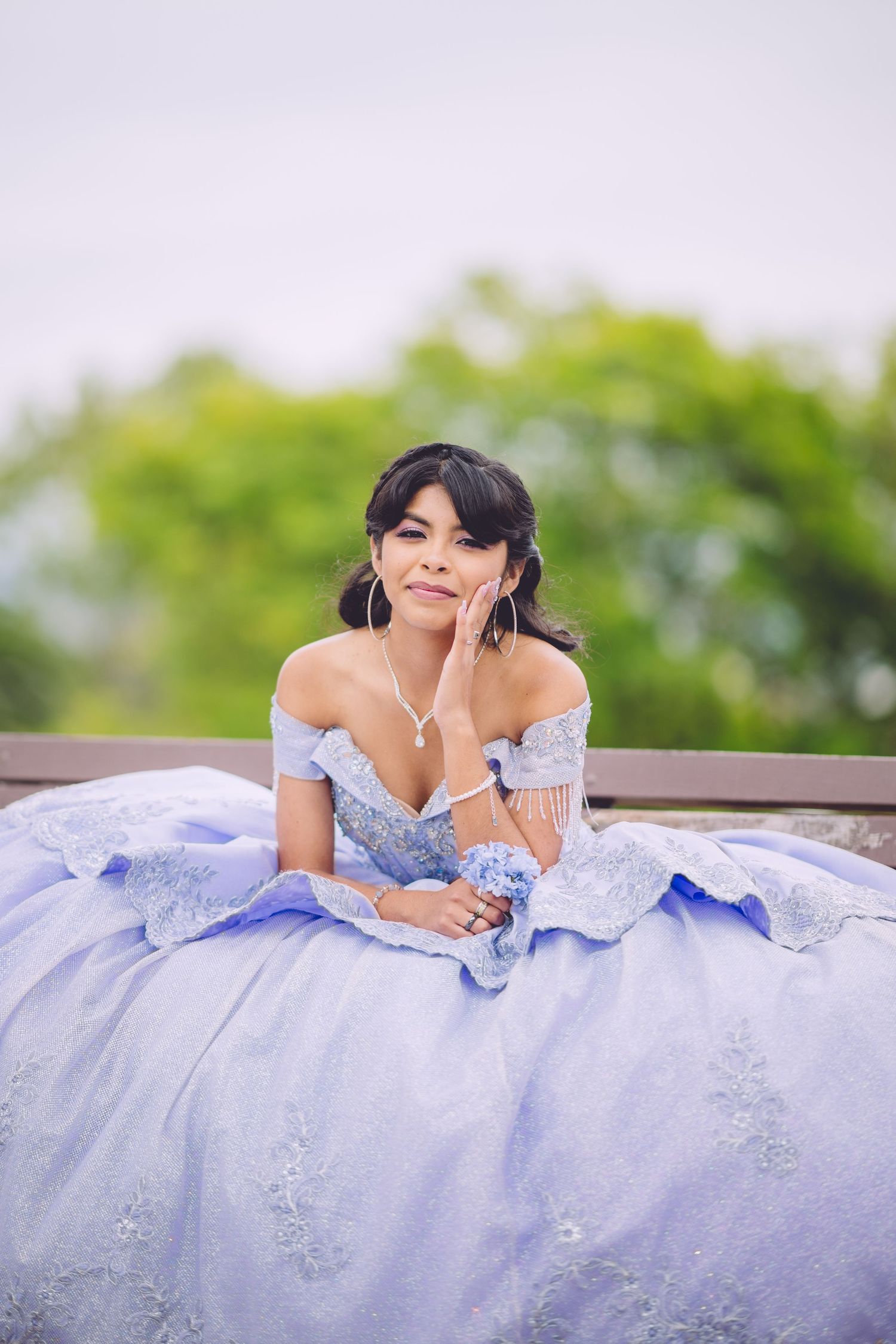 A person in a pale blue off-shoulder ball gown with embellishments poses gracefully outdoors against a blurred green background.