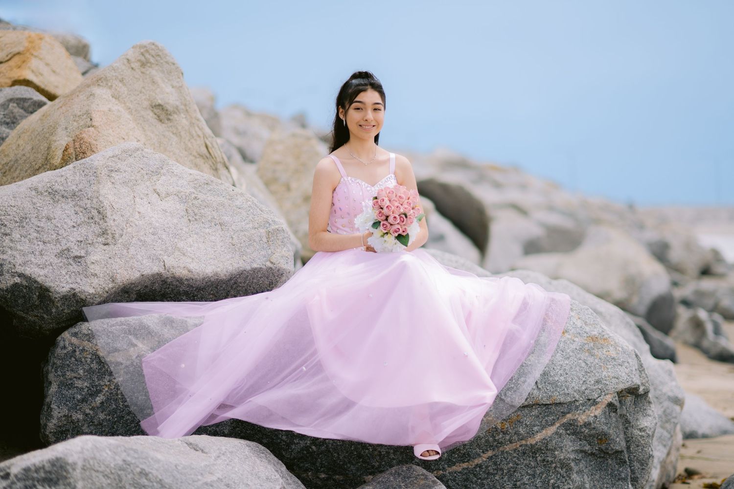 A person in a flowing pink gown poses gracefully on large coastal rocks during a seaside photoshoot.