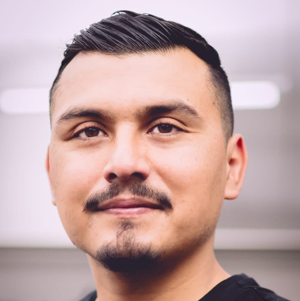 Portrait of person with dark hair styled in an undercut, mustache and goatee, wearing a black shirt against blurred background.