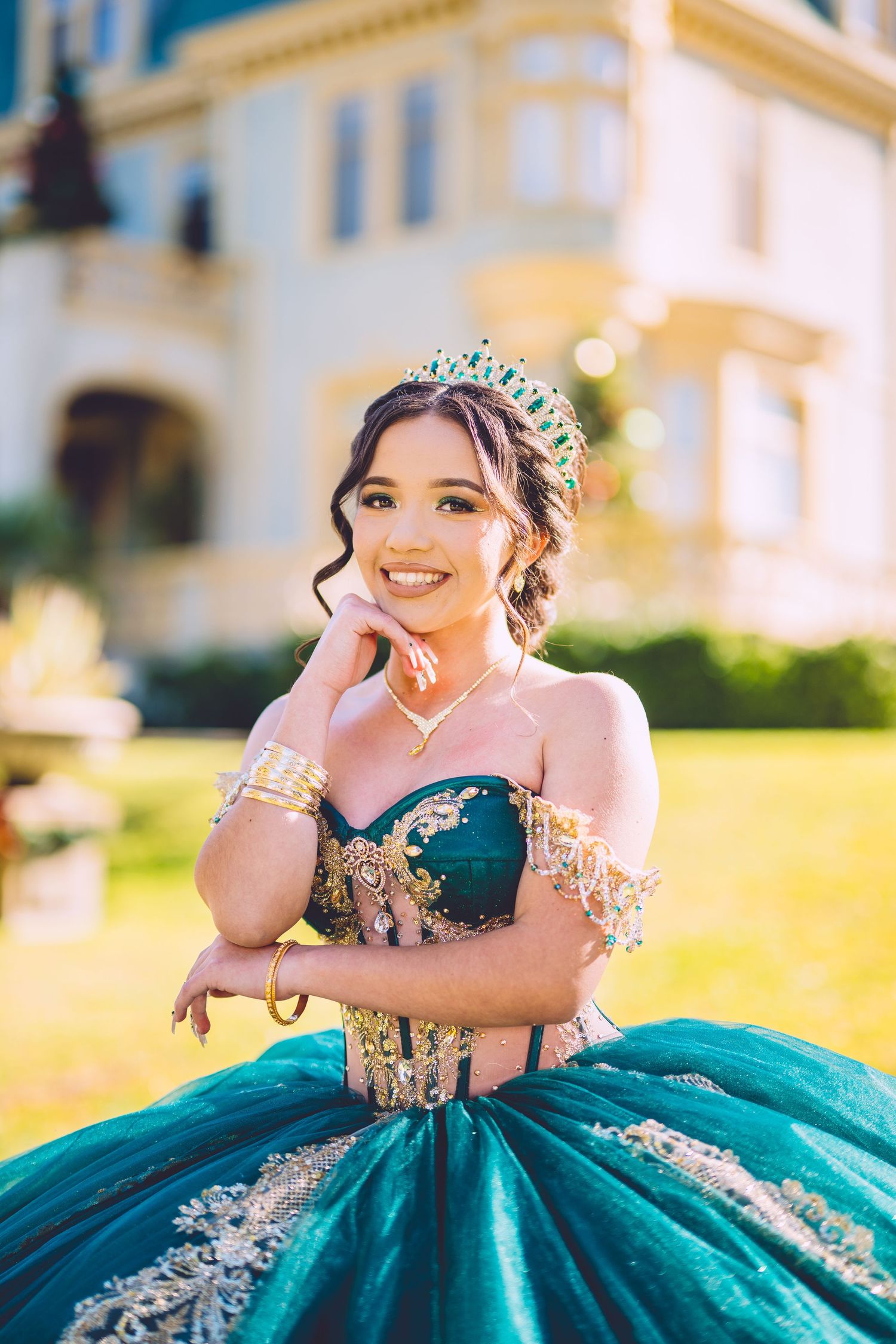 A person wearing an elegant teal and gold quinceañera ball gown with a tiara poses in front of a mansion.