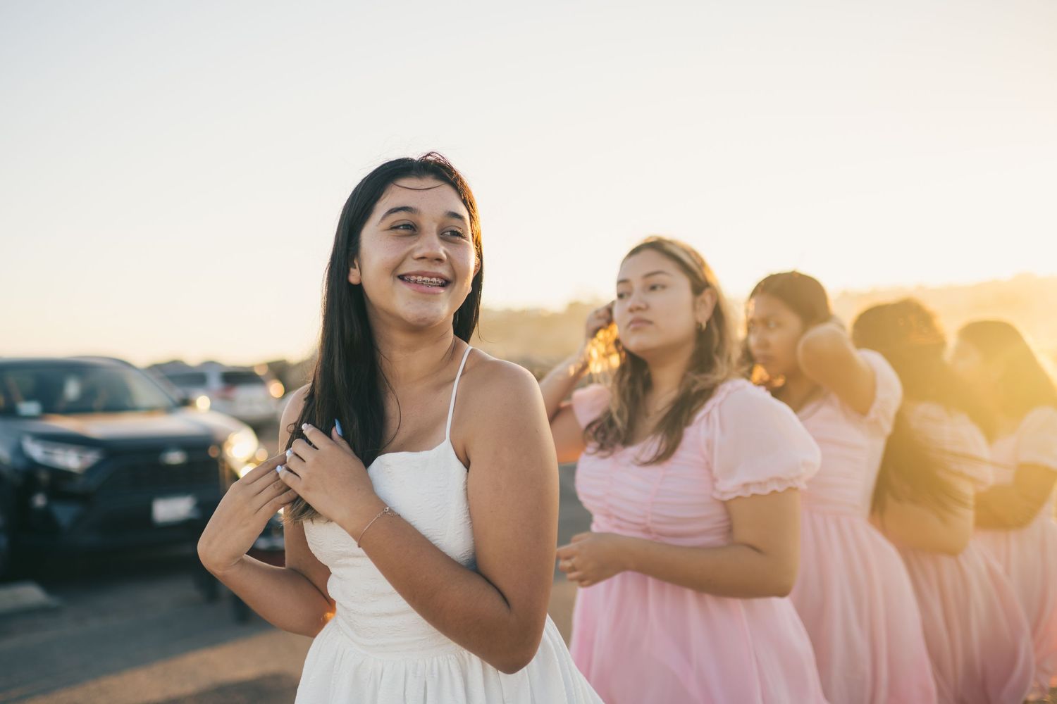 A group wearing white and pink dresses smiles and laughs together at sunset in a parking lot.