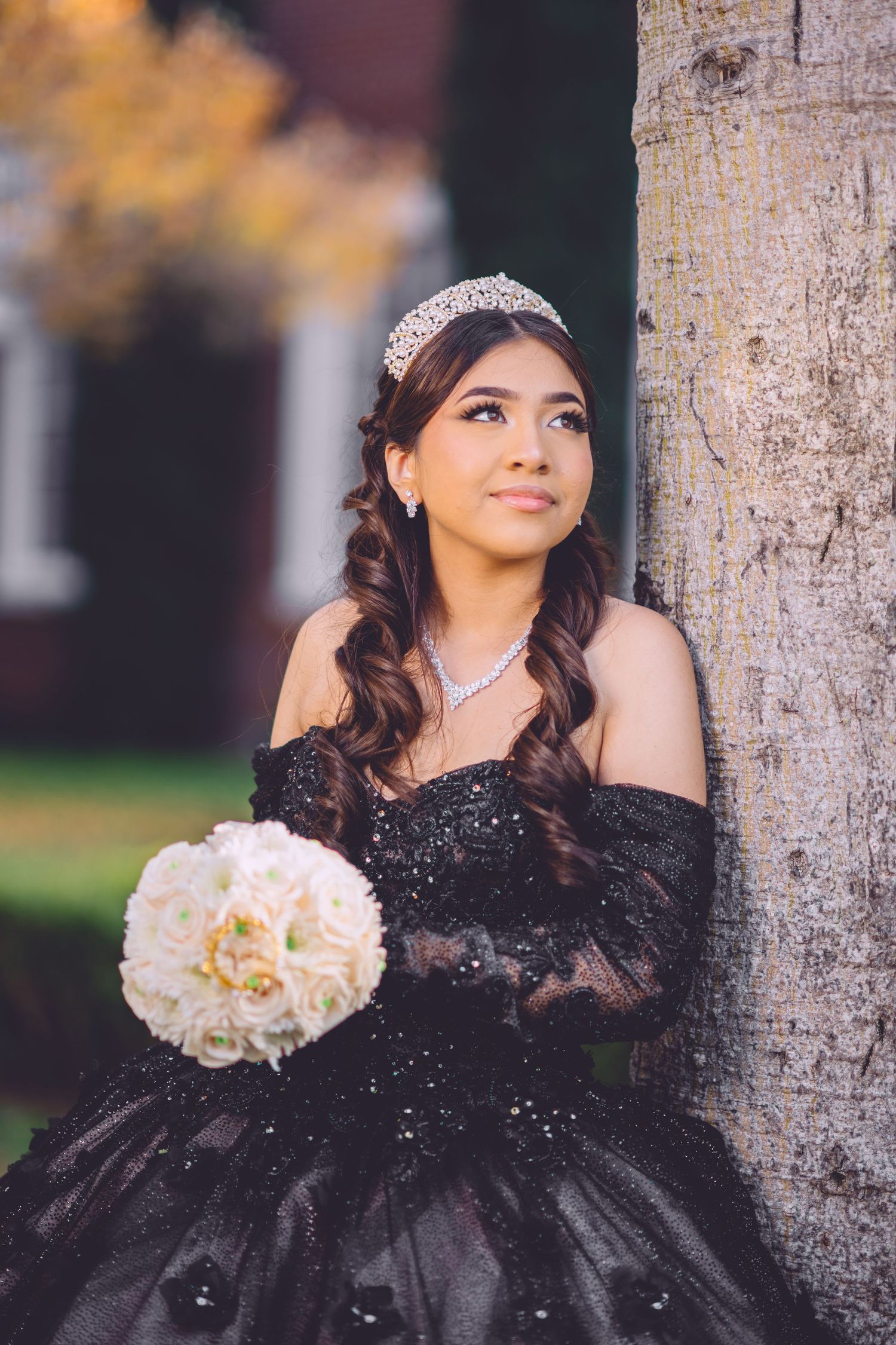 Young woman in black quinceañera gown with tiara holds white bouquet, leaning against tree outdoors.