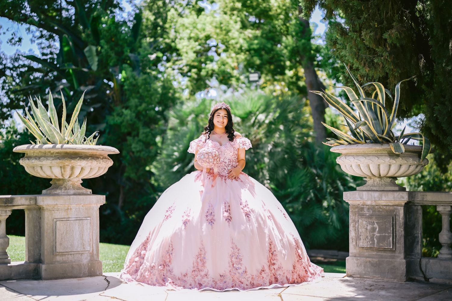 A person in a pink floral quinceanera dress poses elegantly in a garden setting with decorative stone pillars.