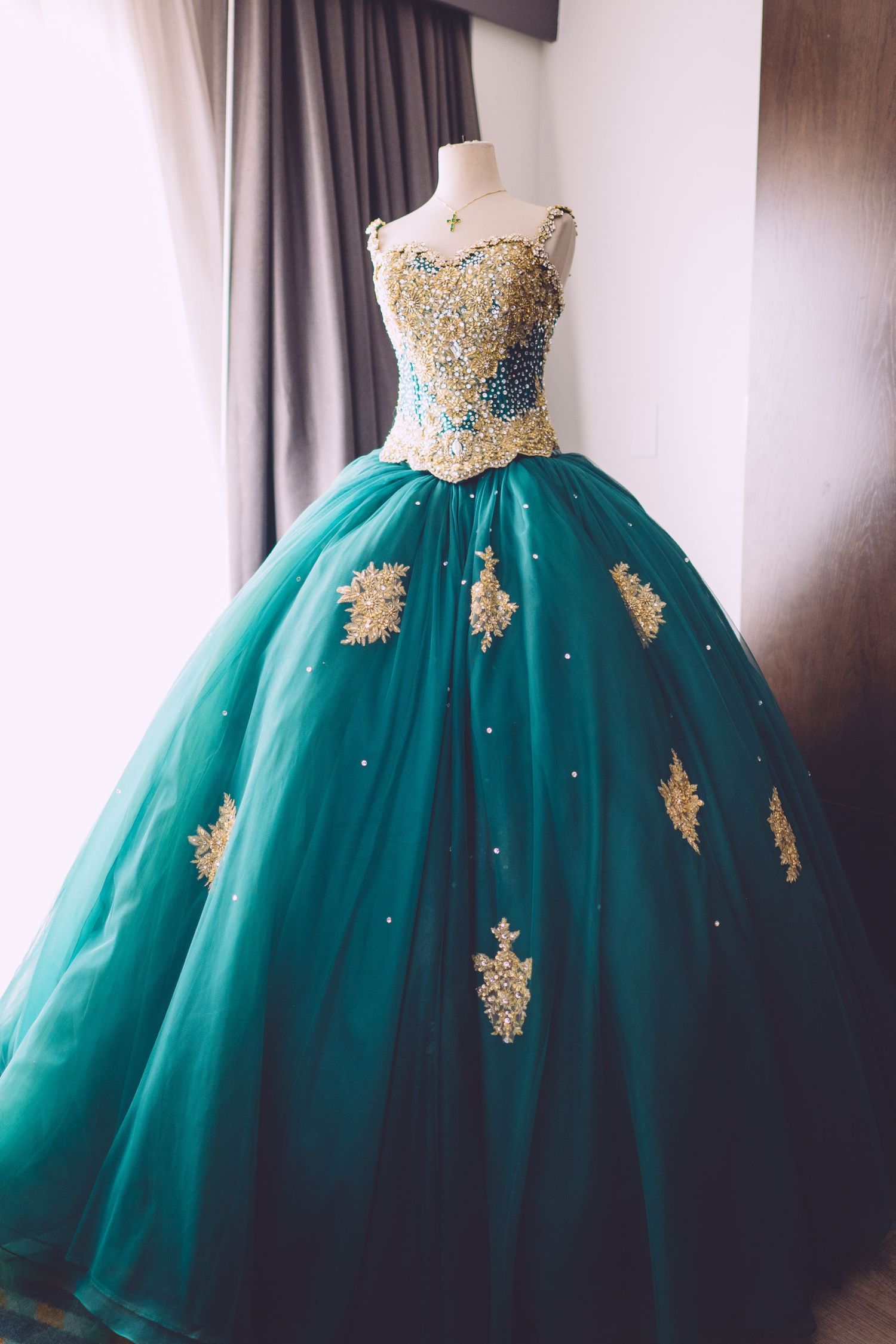 Elegant emerald green ball gown with gold lace appliques and sparkly details displayed on mannequin against curtained wall.