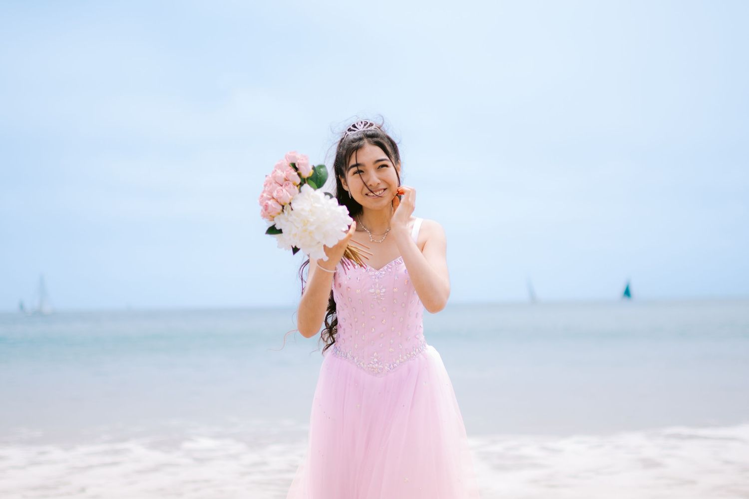 Young woman in pink princess gown and tiara holds floral bouquet on sandy beach, smiling joyfully at camera.