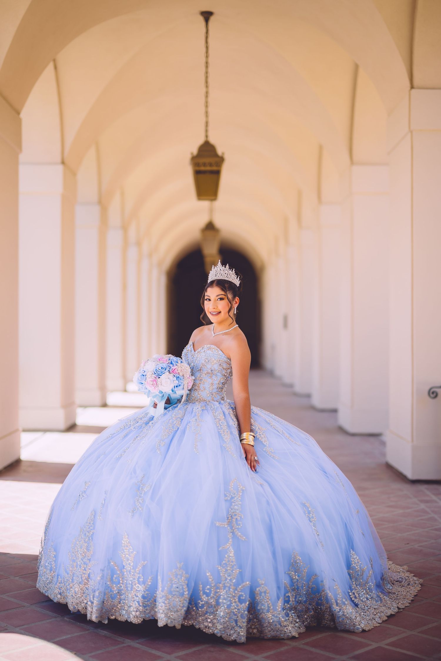 A quinceañera dress in light blue with intricate lace details photographed in an elegant archway corridor.