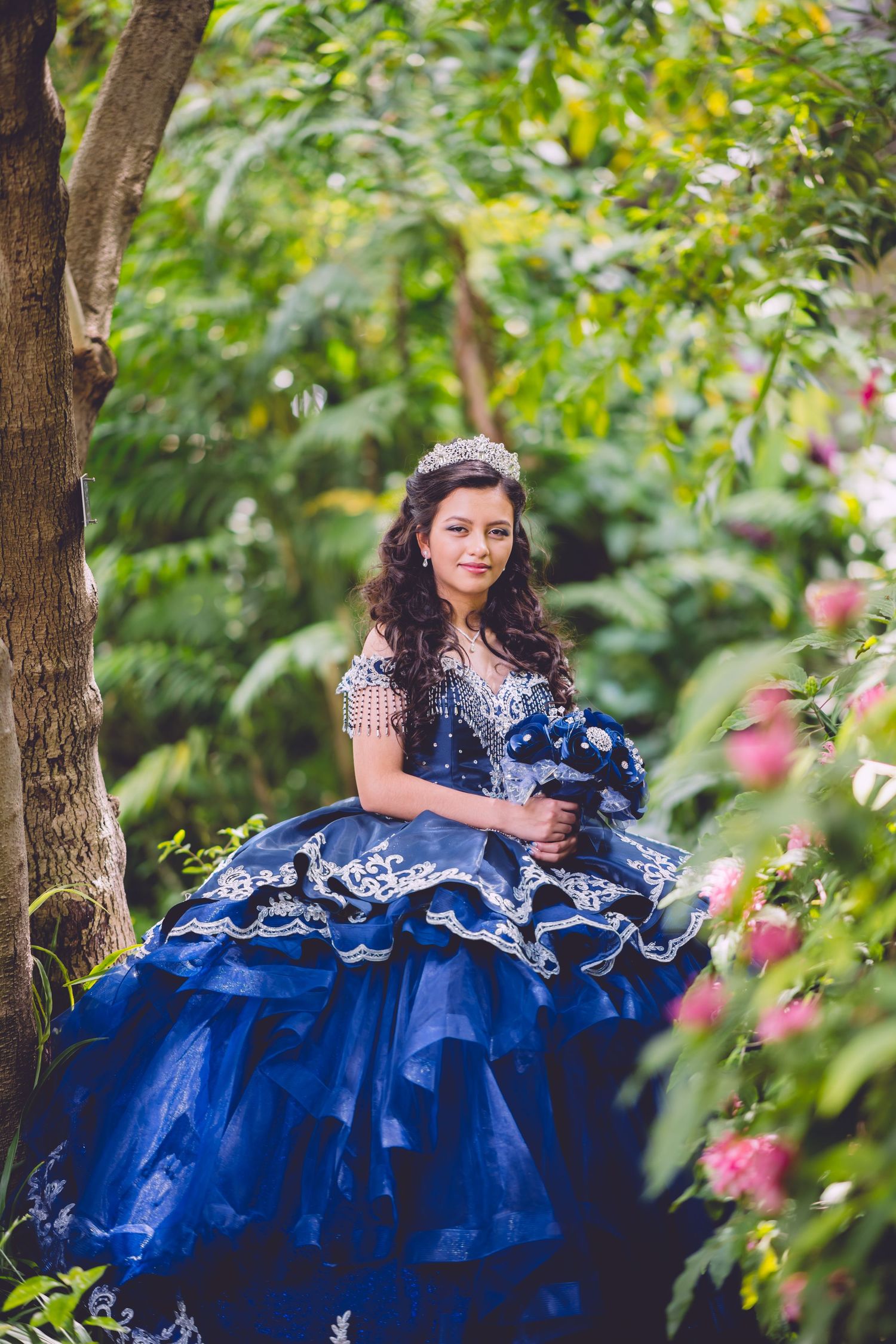 A young person in a royal blue quinceañera dress poses in a garden setting with trees and pink flowers in the background.