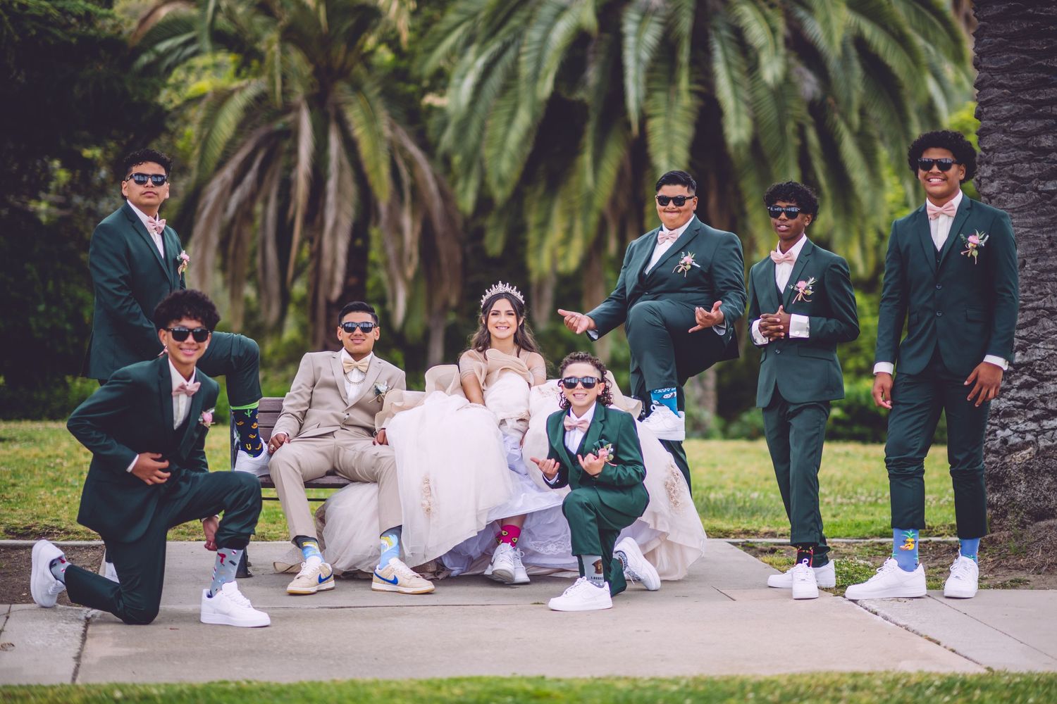 Wedding party in green suits and white sneakers poses casually on grass with palm trees in background.