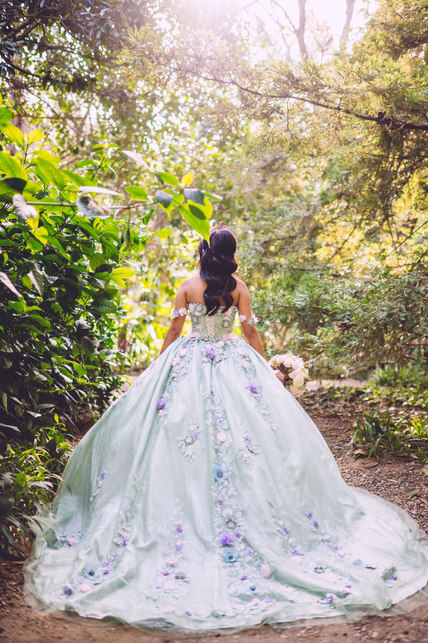 A bride in a white ball gown with purple floral details walks along a sunlit forest path surrounded by lush greenery.