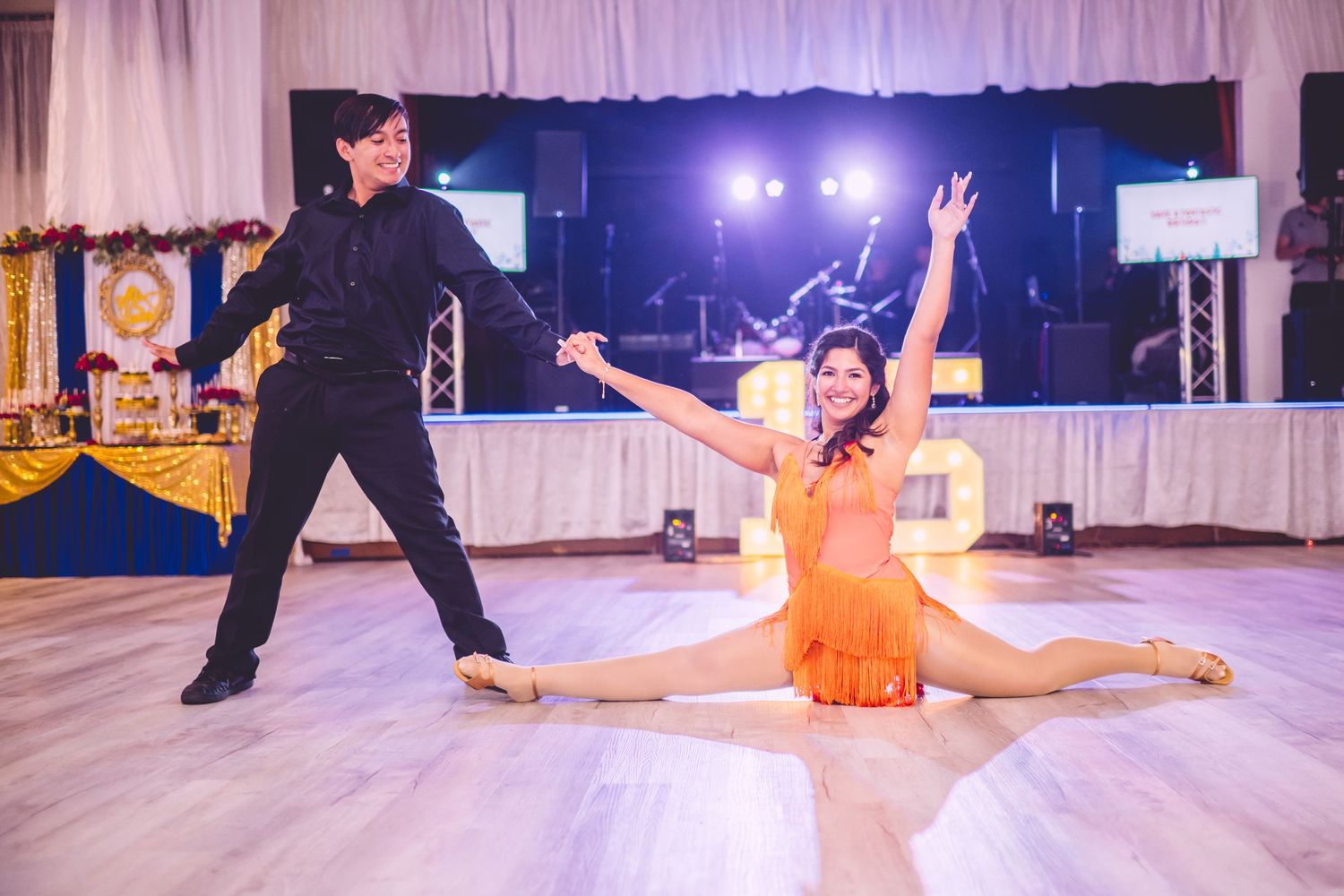 Two dancers perform a dramatic ballroom dance routine with dramatic split pose on illuminated dance floor at event venue.