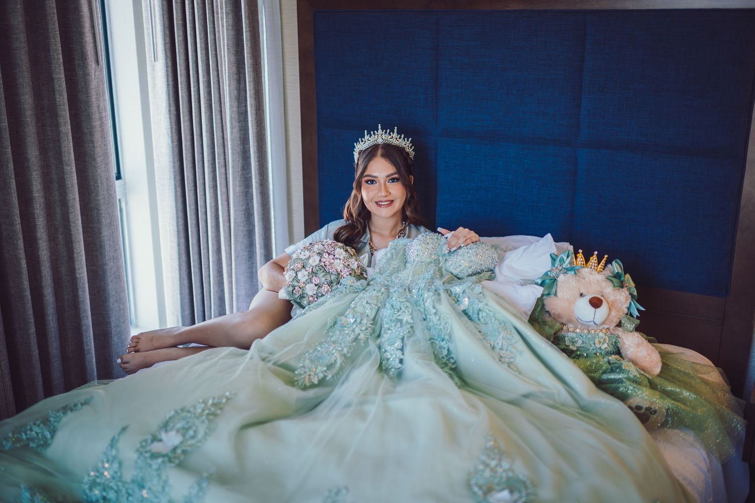 Young woman in mint green quinceañera ball gown with floral crown sits on bed beside a stuffed lion toy.
