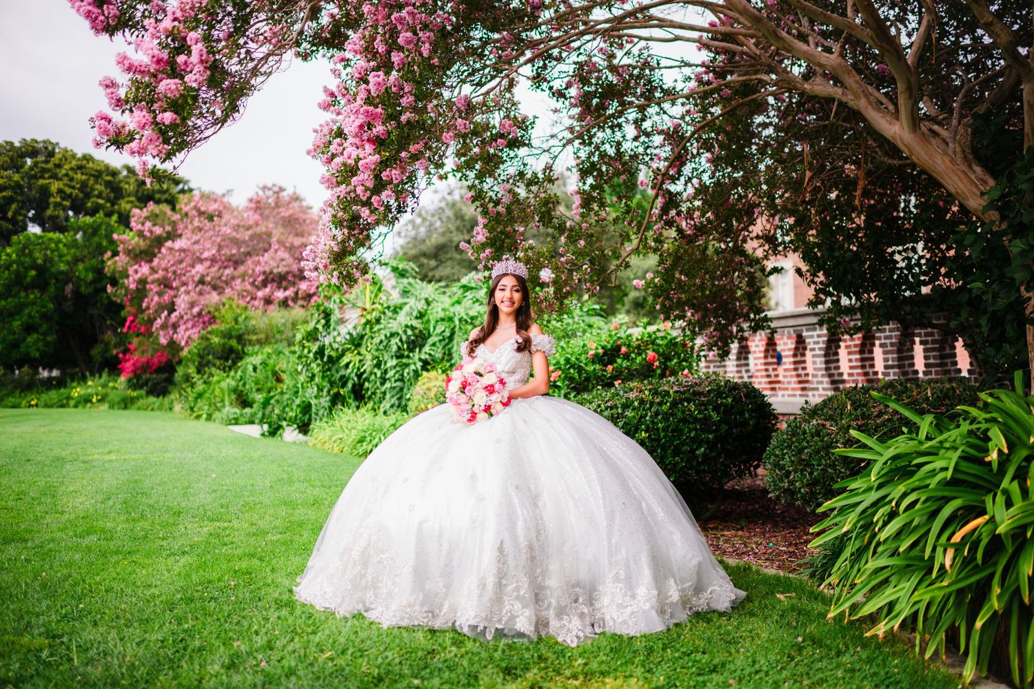 Young woman in white quinceañera ball gown poses in lush garden with pink flowering trees.