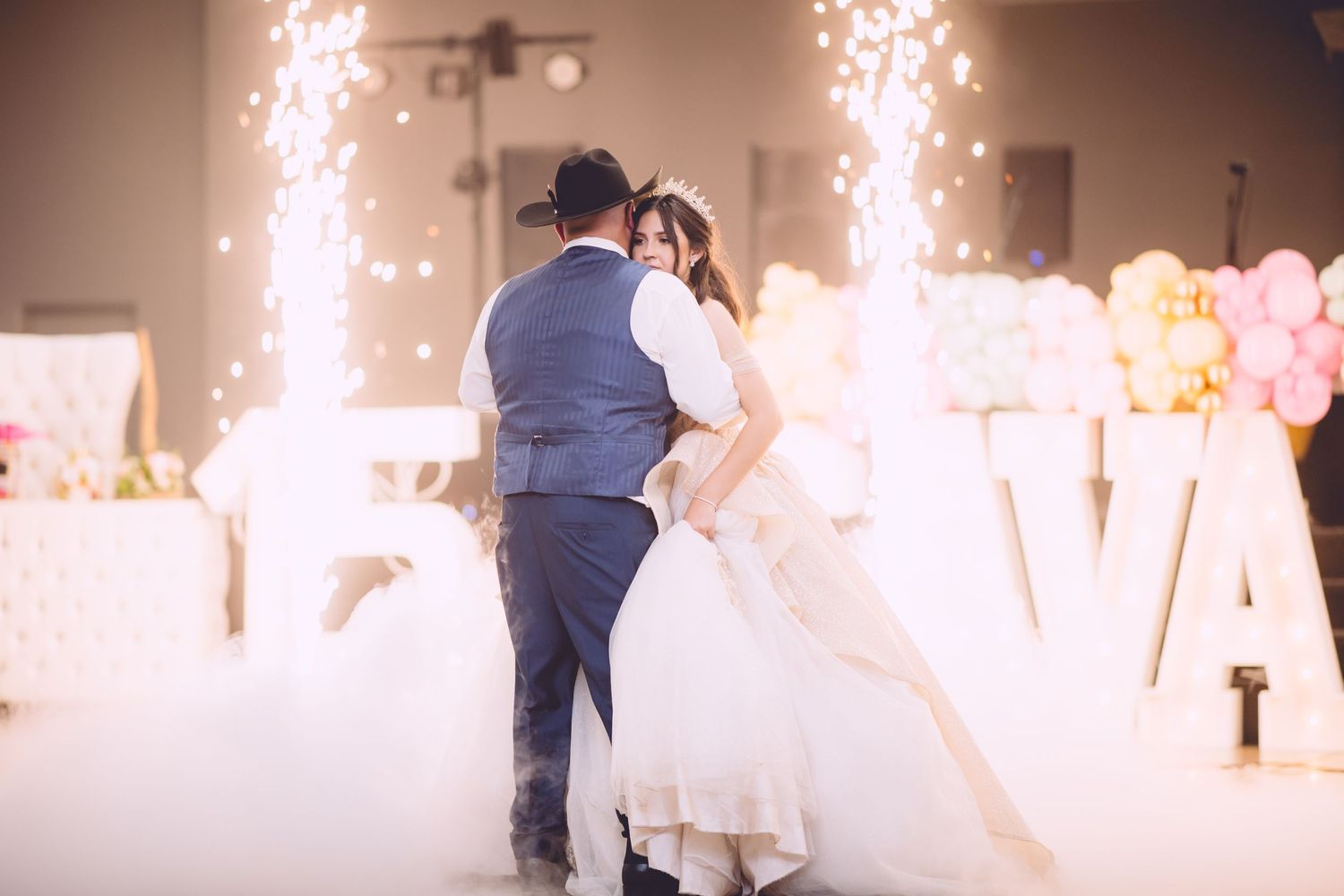 A couple dances their first dance at a wedding reception with sparkling fountains and lit LOVE letters in the background.