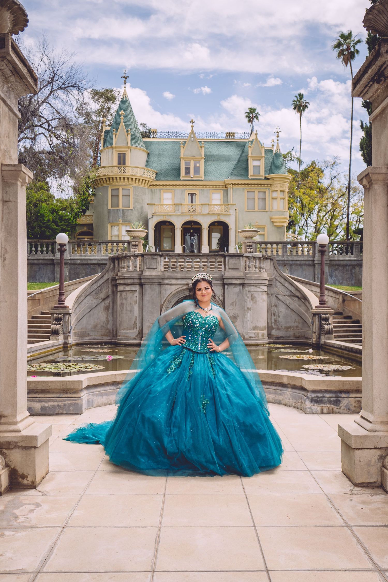 Sparkling teal quinceañera dress showcased at elegant mansion with symmetrical stone staircase.