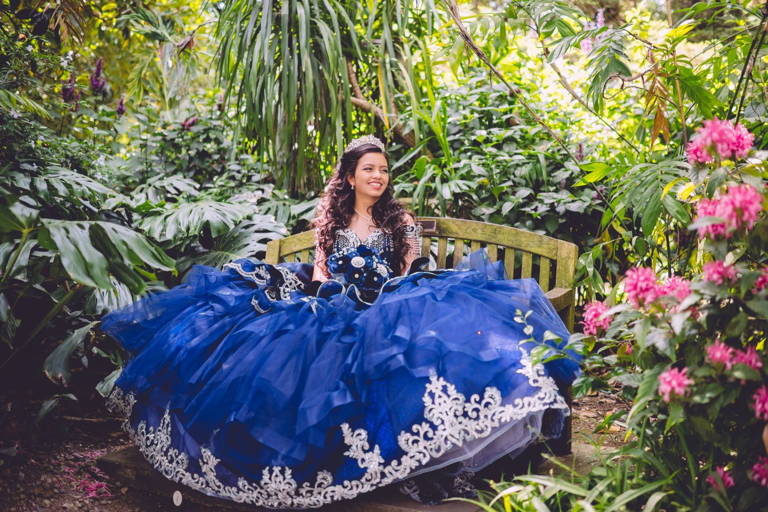 A royal blue quinceañera dress with white lace details photographed in a lush garden setting with pink flowers.