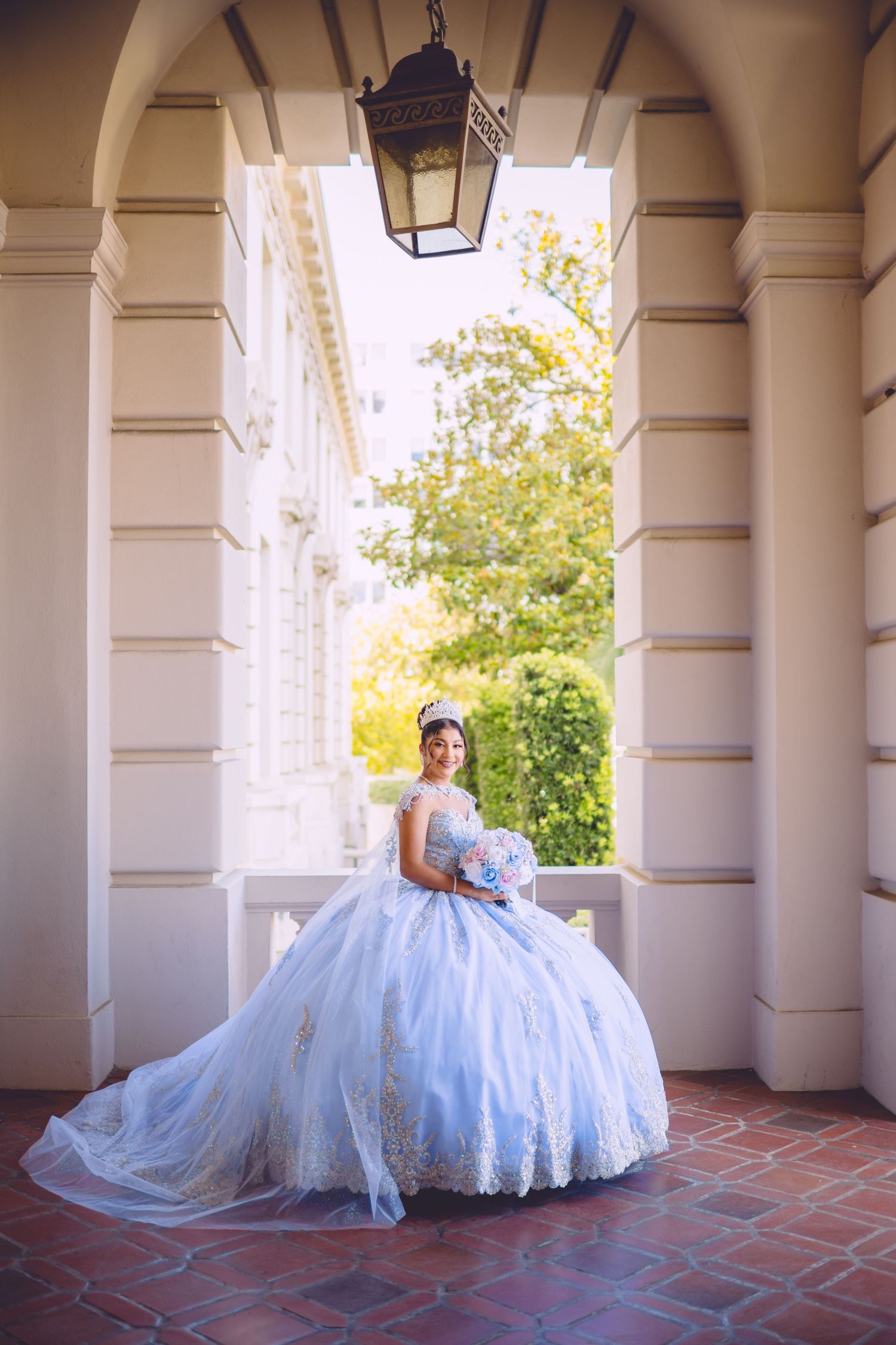 A bride in a voluminous white ball gown poses elegantly under a columned archway on a brick terrace.
