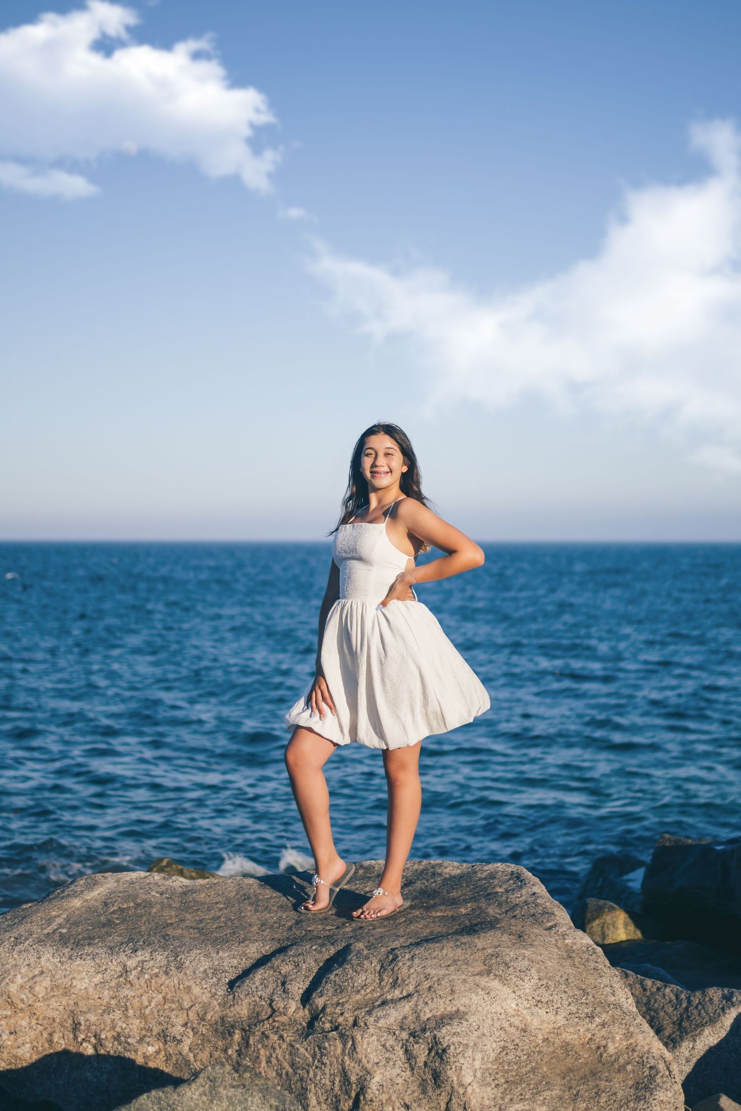 Young woman in white dress standing barefoot on rocks by the blue ocean under a partly cloudy sky.
