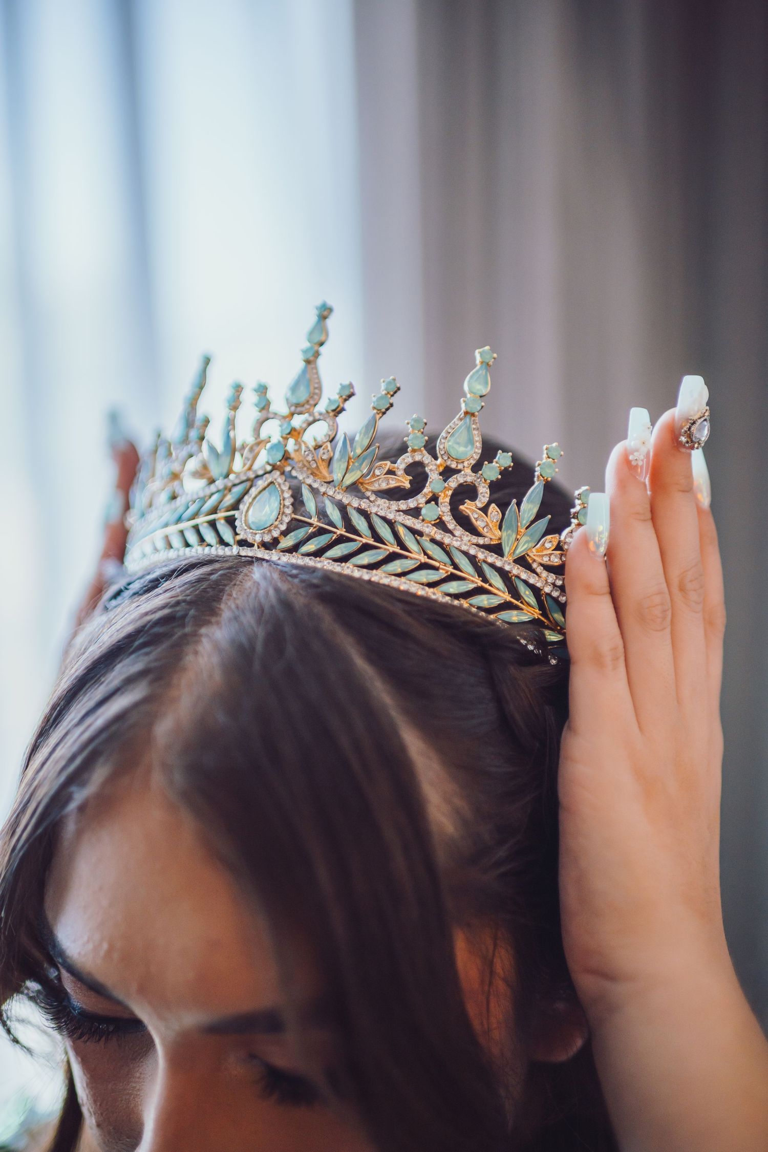 Woman placing an ornate blue and gold tiara crown on dark hair, with manicured white nails visible.