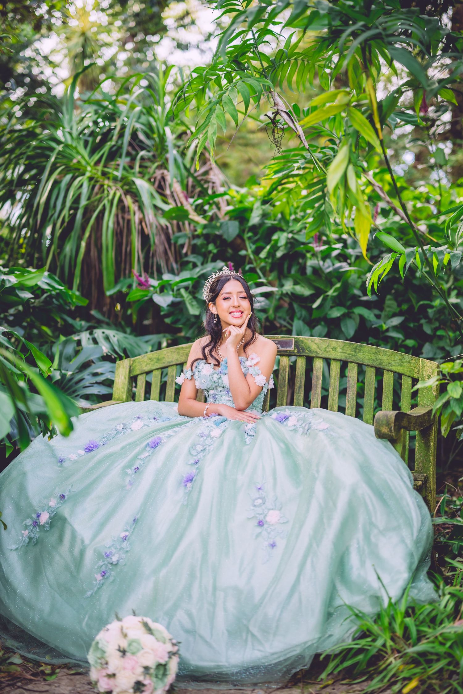 A person in a light blue ballgown with floral details sits on a wooden garden bench surrounded by lush tropical foliage.
